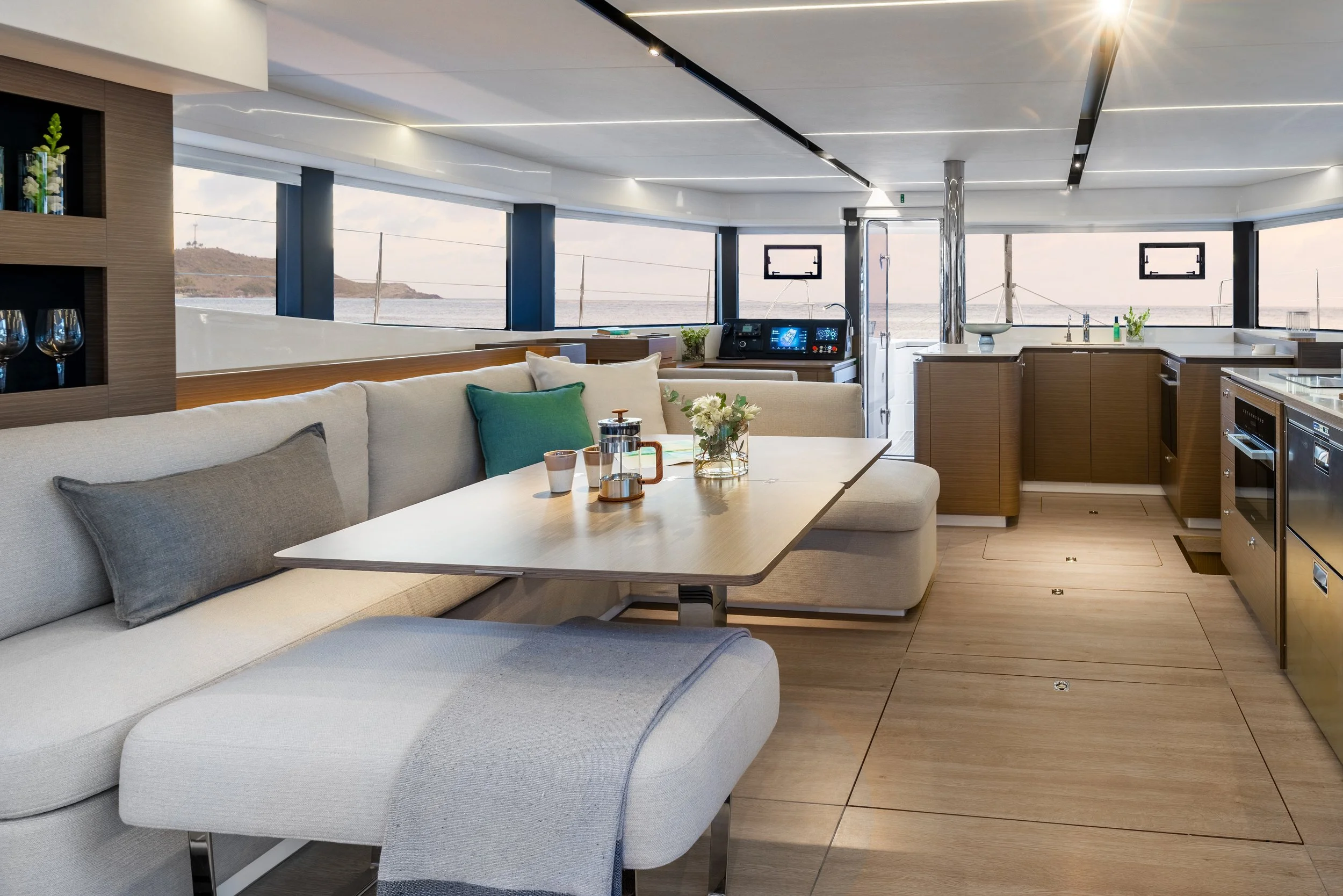 Interior view of a modern yacht's living area with a view of the ocean outside the large windows, including a white sofa, a dining table with cups and a French press, and a kitchen area with wooden cabinets and appliances.