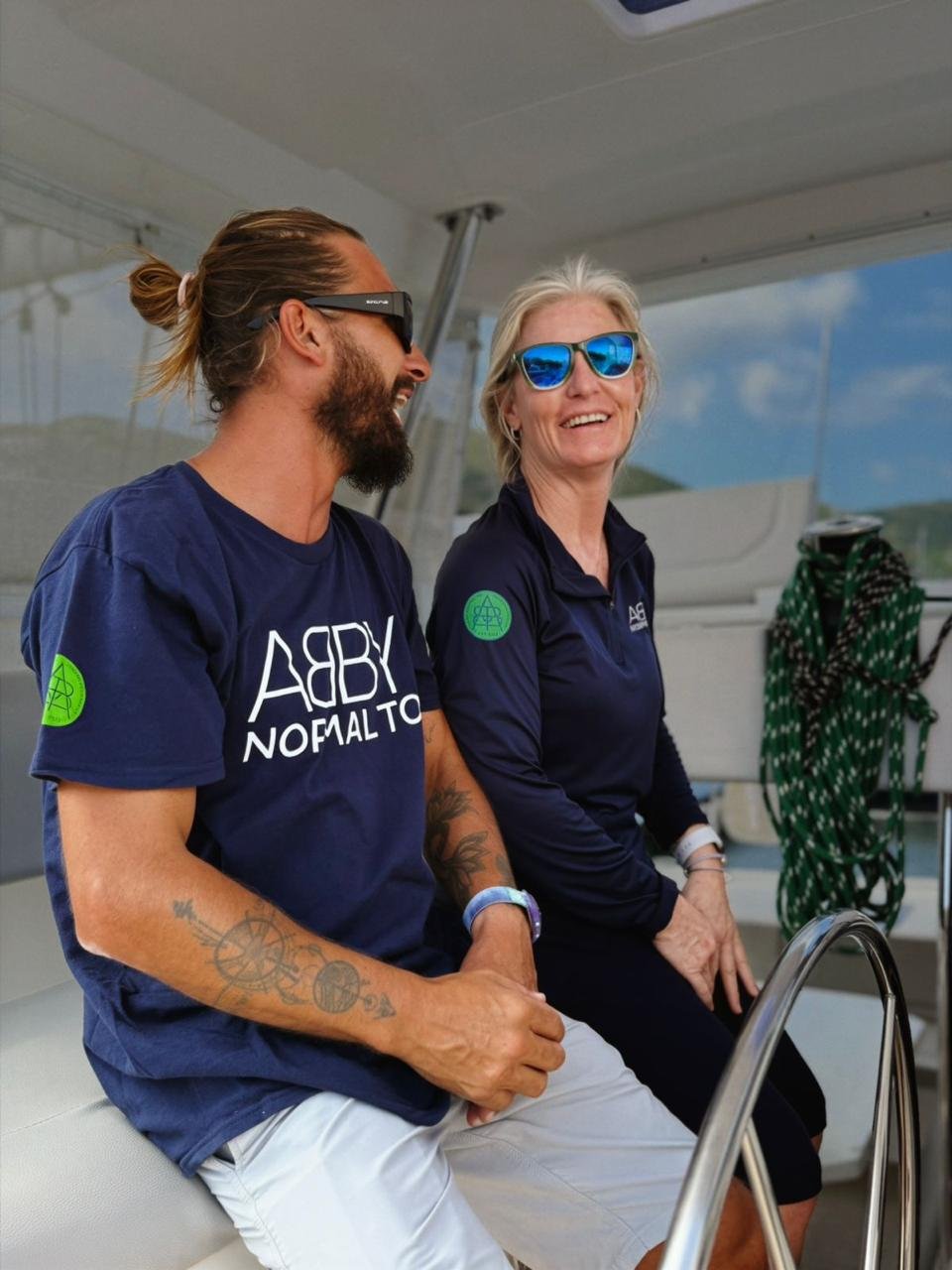 Two people sitting on a boat, smiling and wearing sunglasses, with a background of blue sky and water.