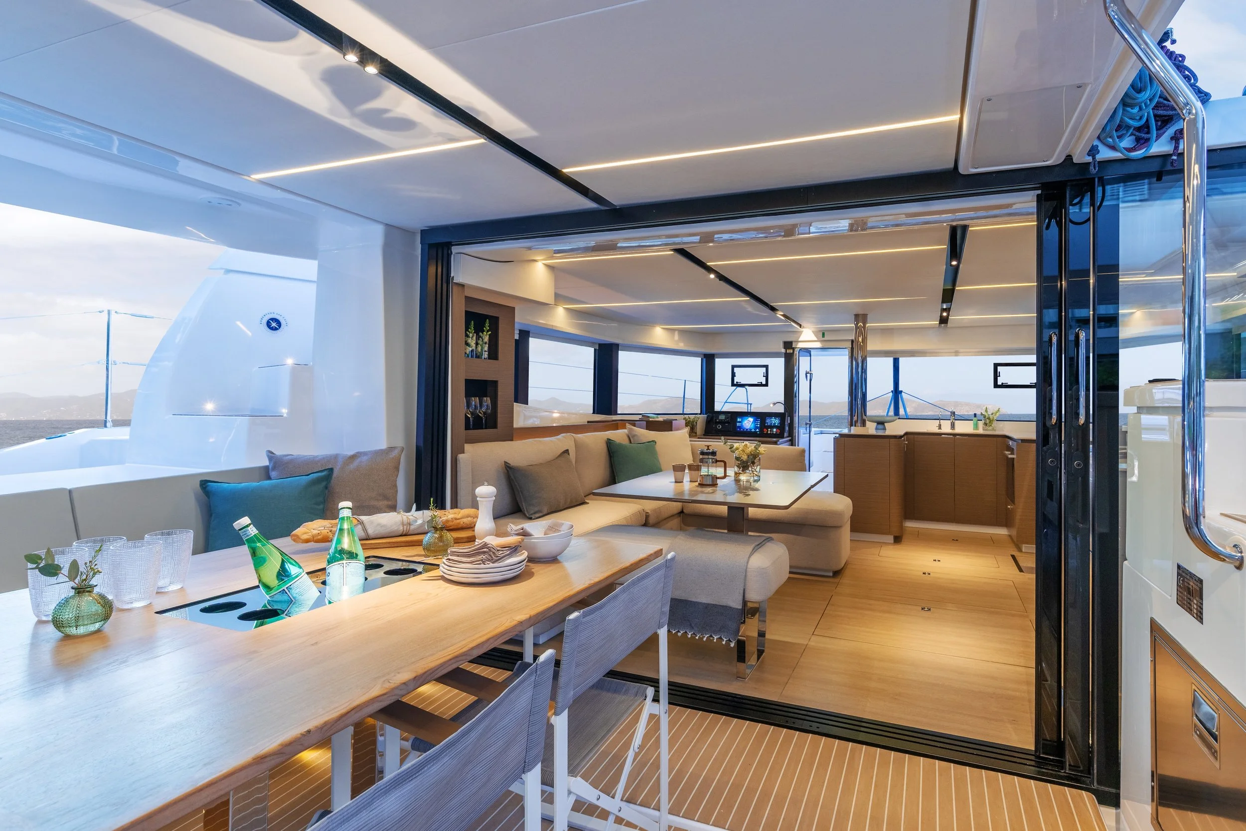A modern yacht outdoor dining area and a seating lounge, featuring a wooden table set with glasses and bottles, a beige sofa with cushions, and large windows connecting the indoor and outdoor views.