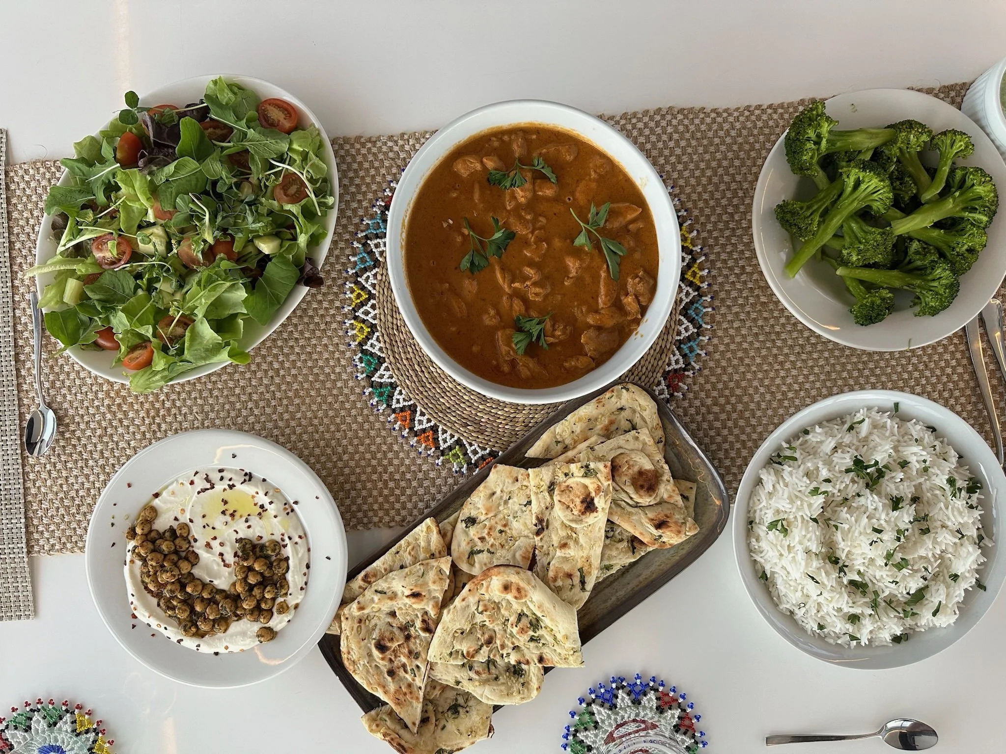 A table with a variety of Indian foods including a bowl of curry, naan bread, rice, a side salad, and yogurt with chickpeas, served on a woven placemat.