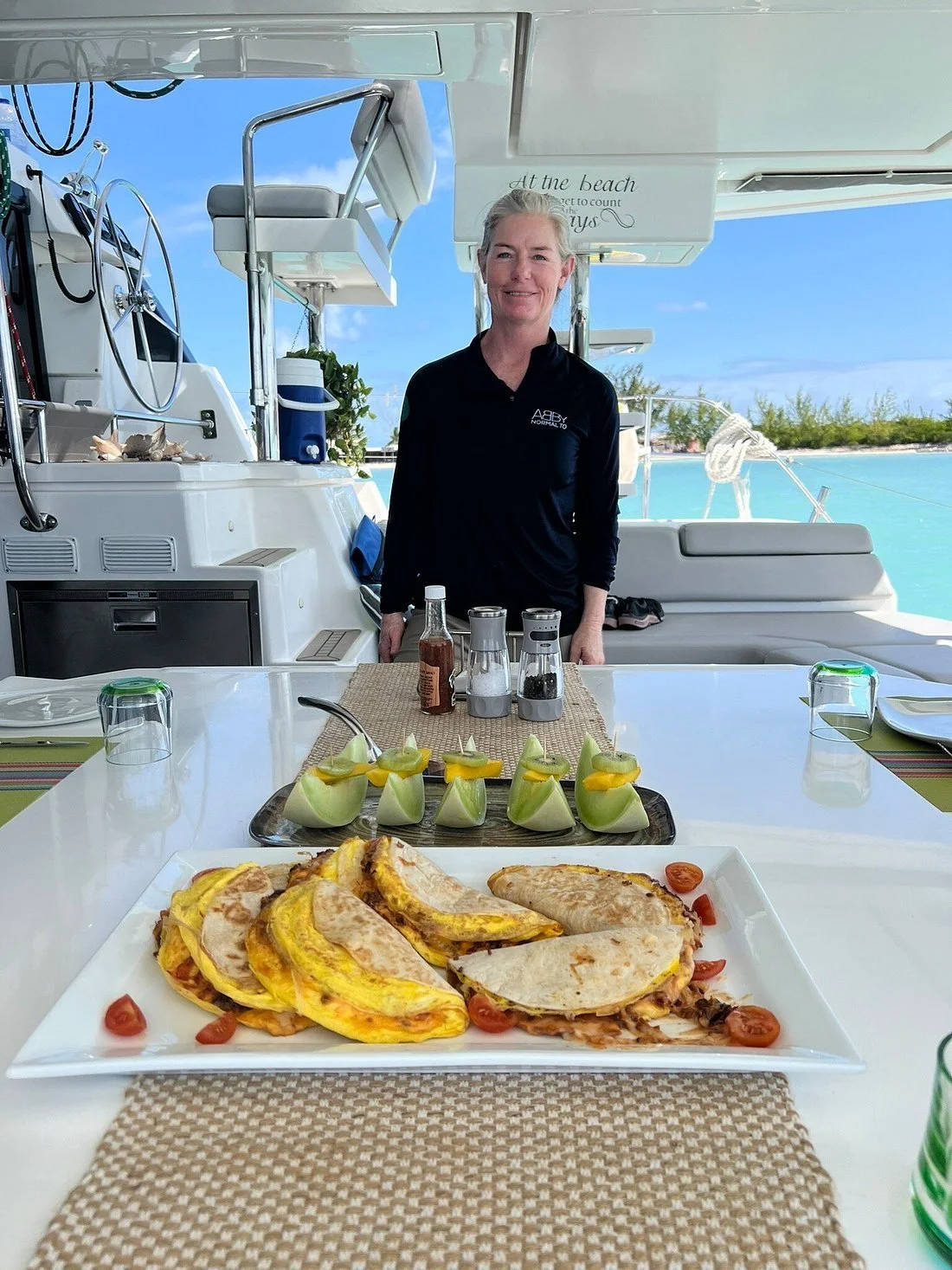 A woman standing behind a table on a boat, with plates of food including quesadillas and sliced vegetables, set for dining near the ocean with a blue sky in the background.