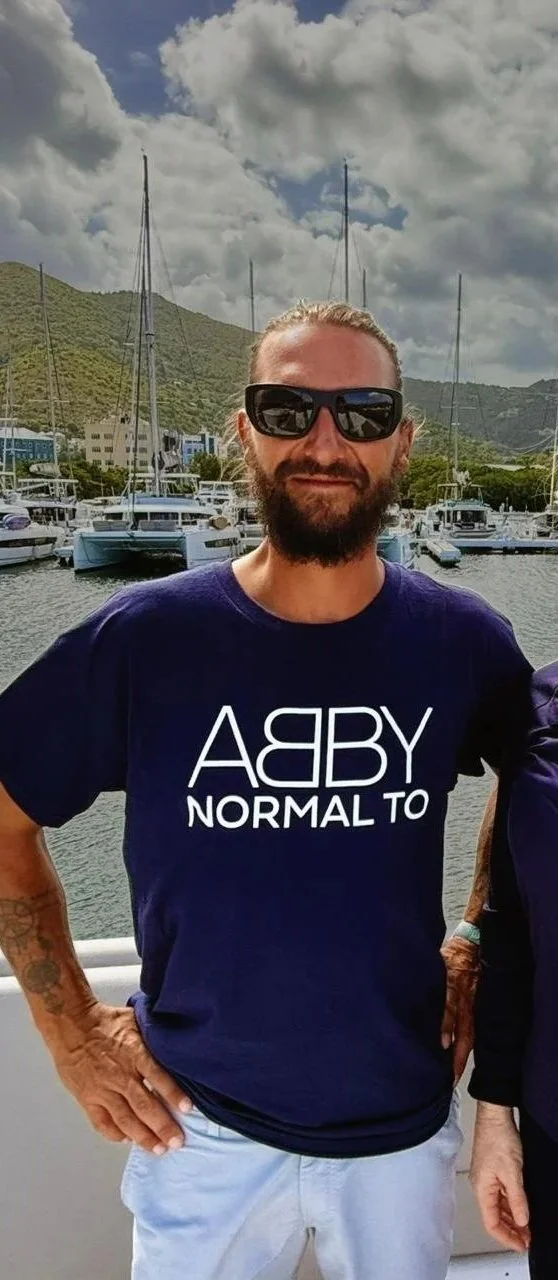 A man with a beard and sunglasses standing outdoors by a marina with sailboats and mountains in the background, wearing a navy T-shirt that says 'YABBA NORMAL TO'.