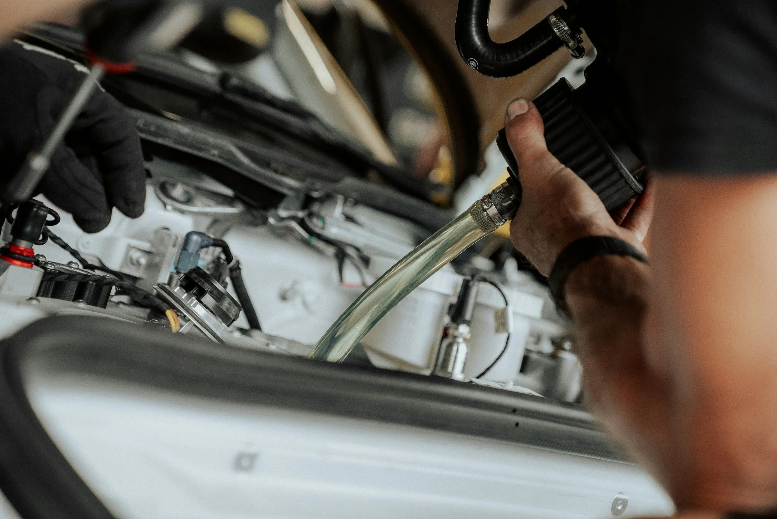 Person working on a car engine, holding a black component with their right hand, amidst various engine parts and tools.