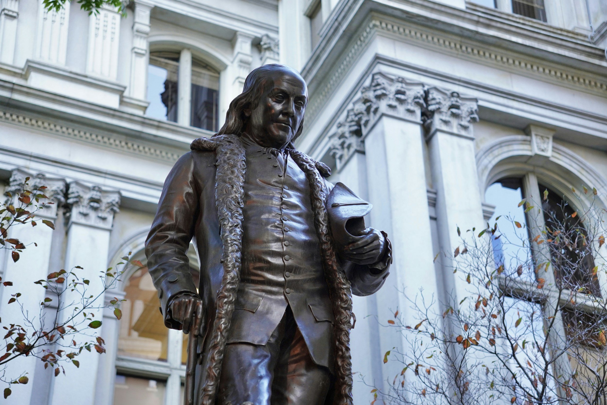 Bronze statue of Benjamin Franklin outside a historic building with ornate architecture.