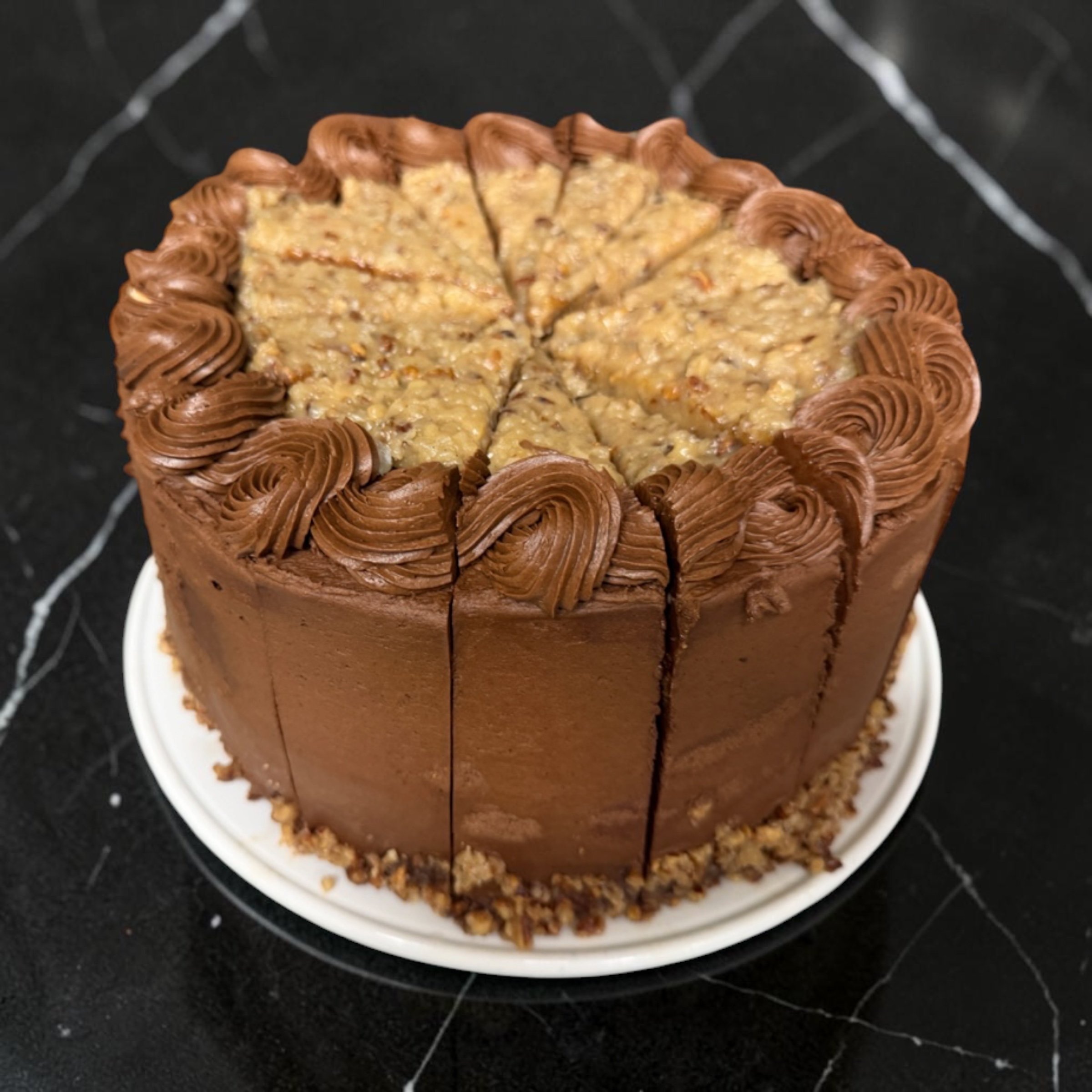 A tall, round cake with chocolate frosting and decorated with piped chocolate swirls, topped with a layer of crushed nuts.