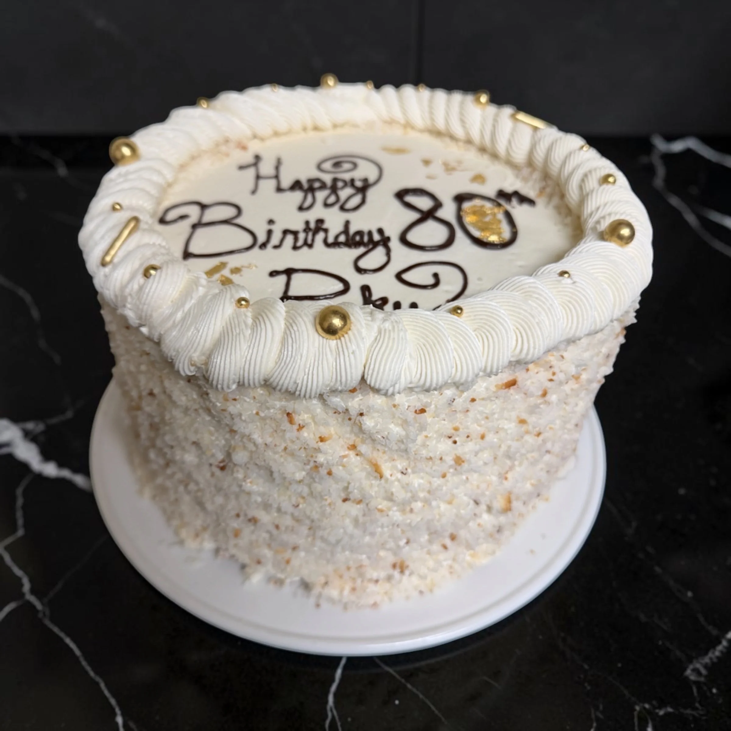 A round birthday cake with white frosting, decorated with gold and white spherical accents around the top edge, and writing that says "Happy 80th Birthday" in black icing.