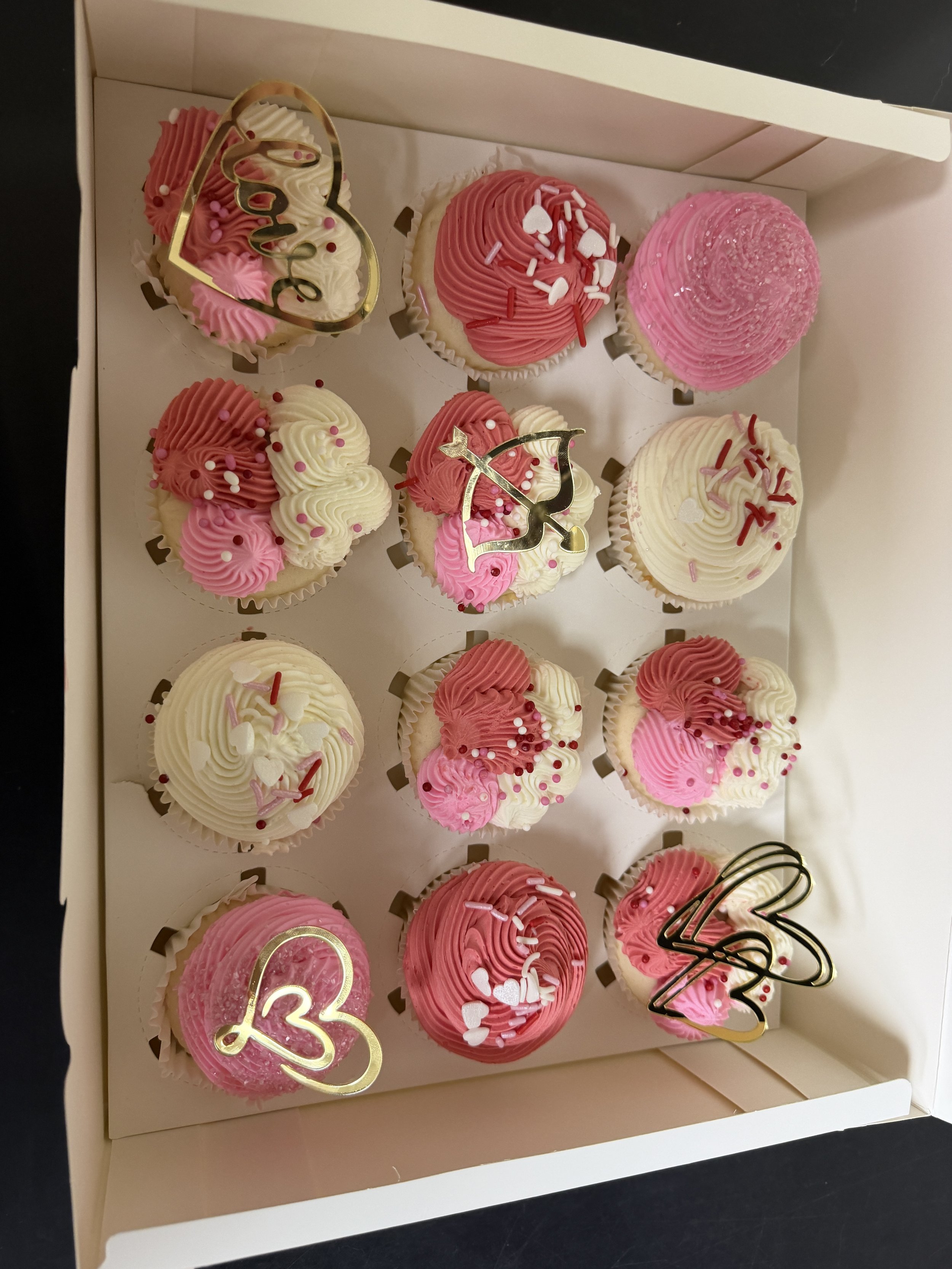 A box of pink and white cupcakes decorated with sprinkles, face-shaped toppers, and a number '13' topper, indicating a birthday celebration.
