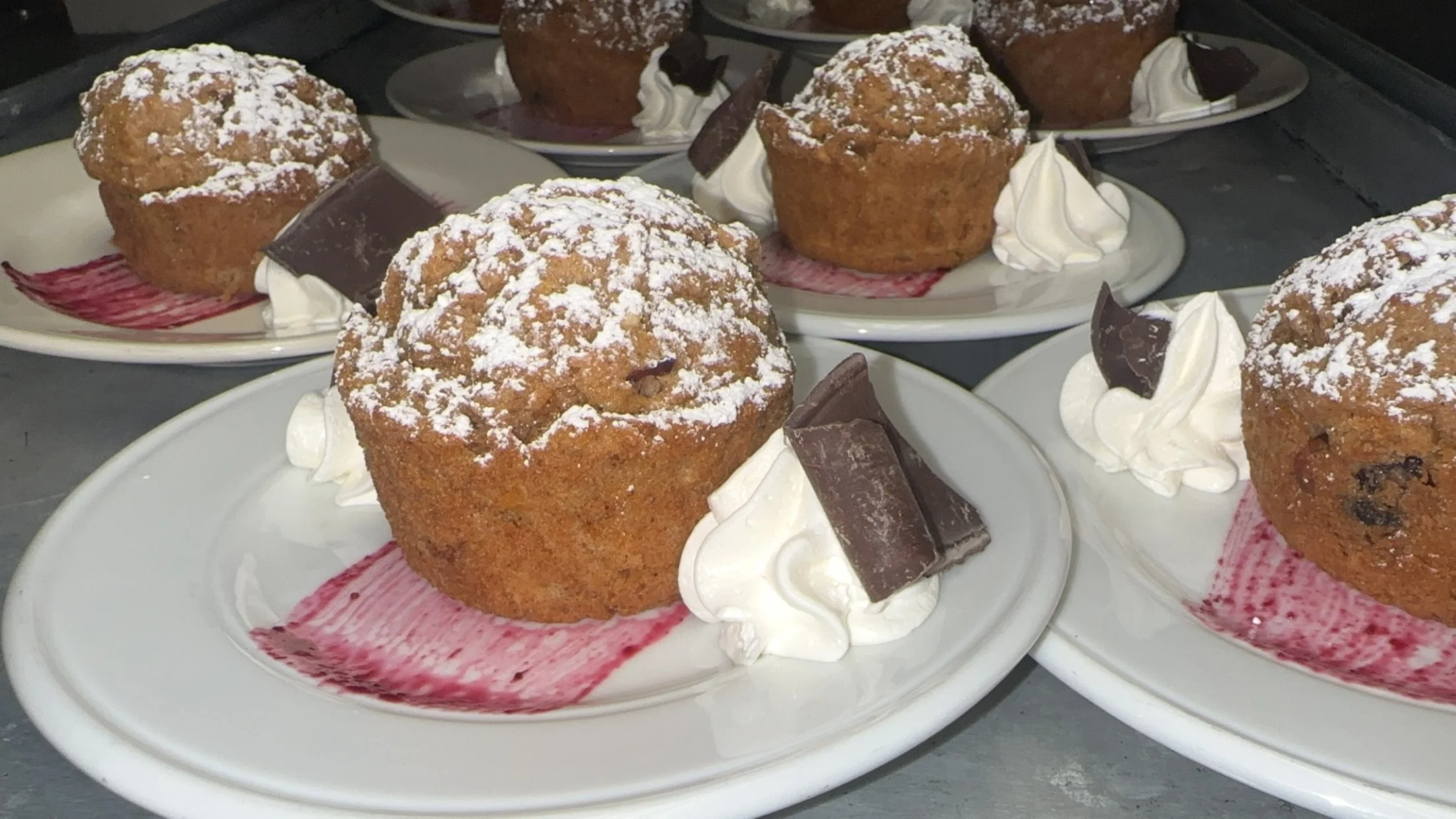 Several plates of blueberry muffins garnished with powdered sugar, nested among dollops of whipped cream and pieces of chocolate, on a dark surface.