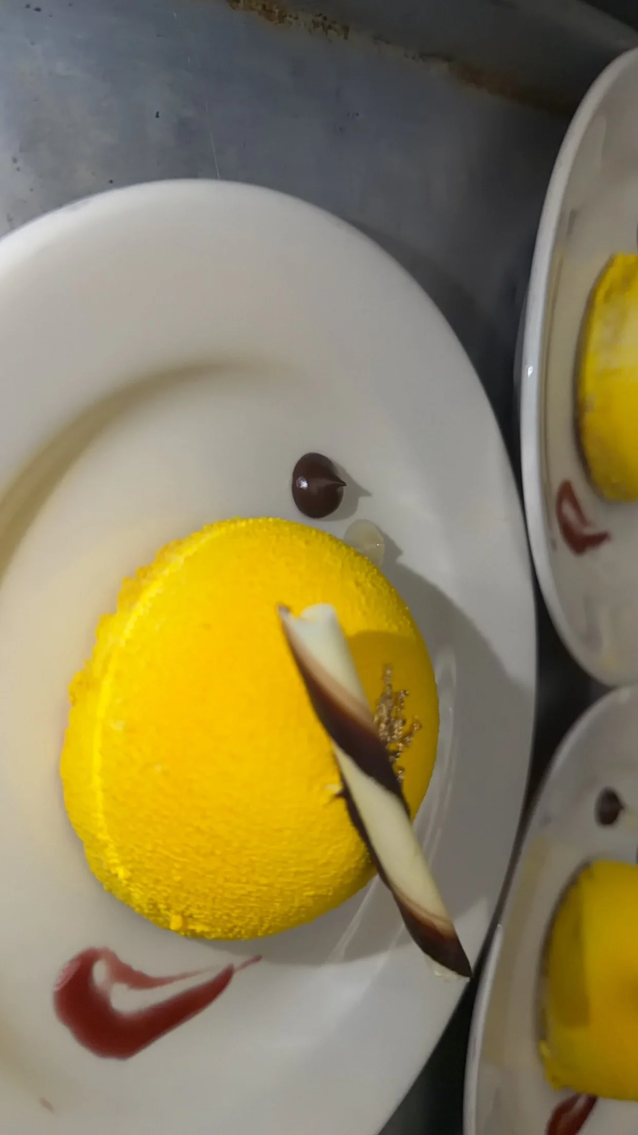 A yellow cake with a smooth iced surface on a white plate, decorated with a strip of white chocolate and a dark chocolate curl, with chocolate drizzle on the plate background.
