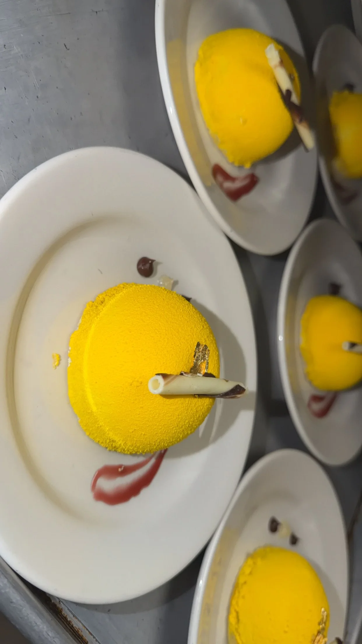 A yellow domed dessert on a white plate garnished with a white chocolate curl, served with a drizzle of red syrup.