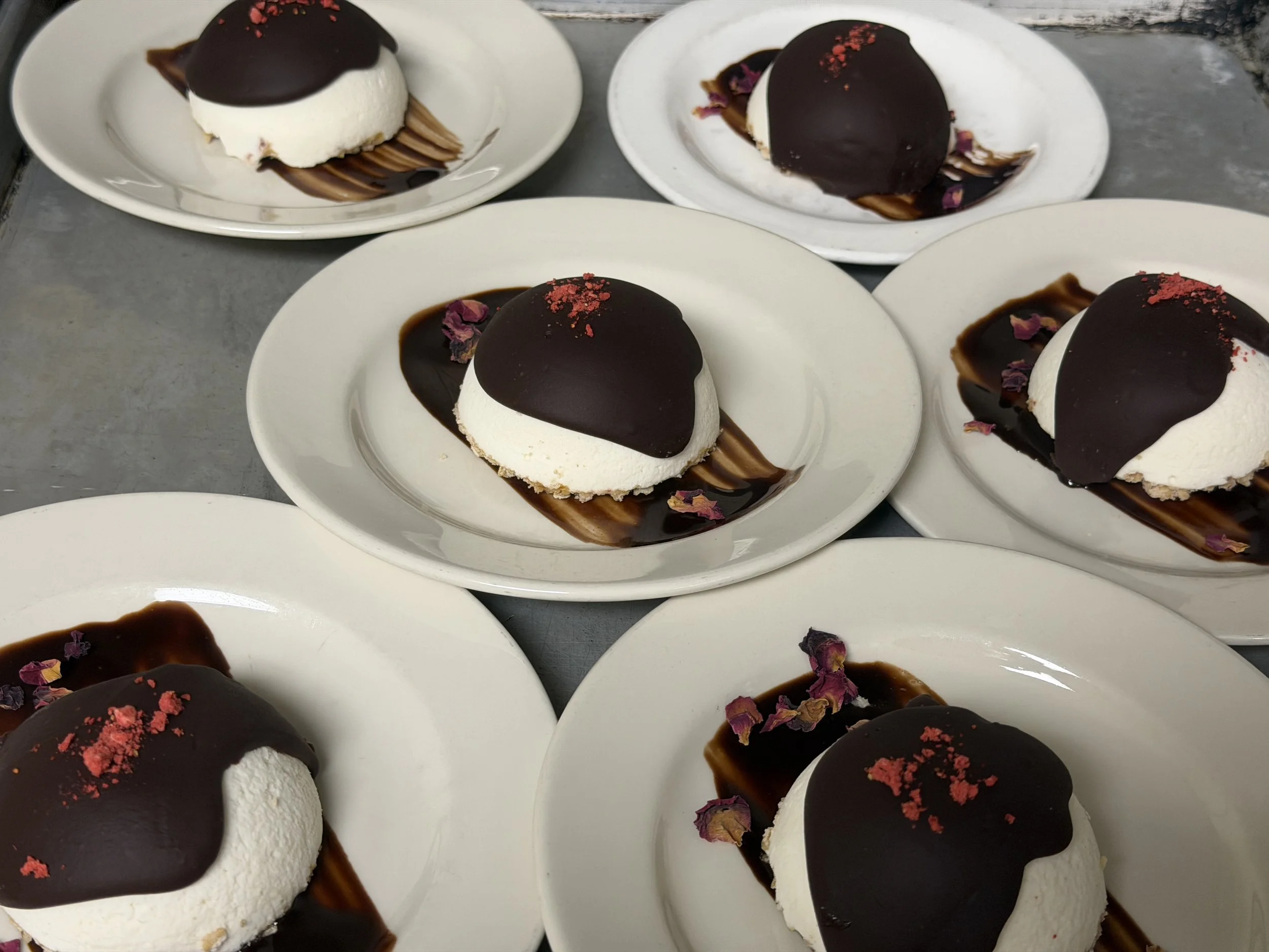 Plate of individual desserts with white cream, topped with glossy chocolate coating, sprinkled with red powder and dried rose petals, garnished with chocolate sauce underneath.