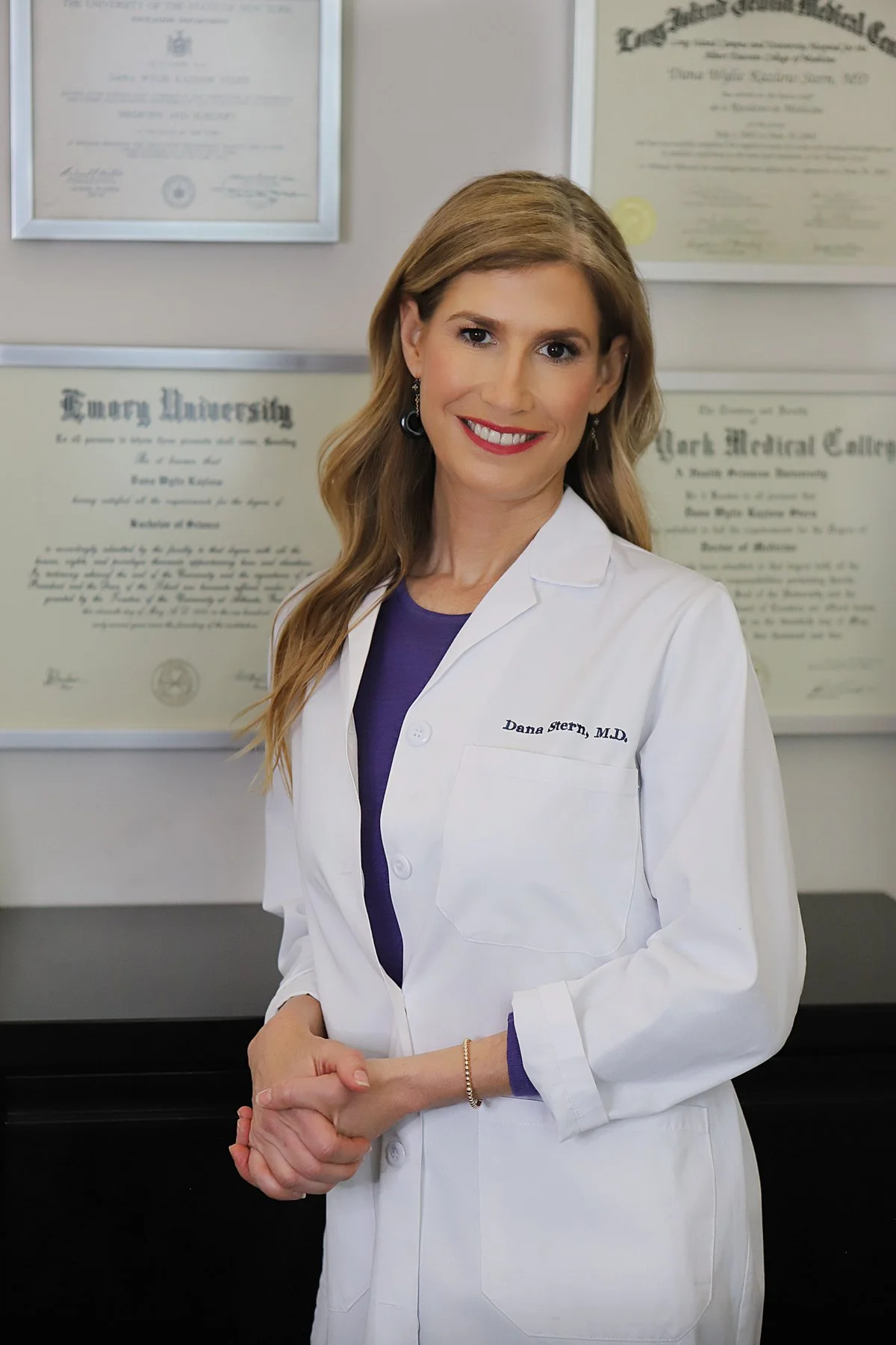 A woman smiling in a white lab coat standing in front of framed medical diplomas, with her hands clasped in front of her, wearing earrings and a bracelet.