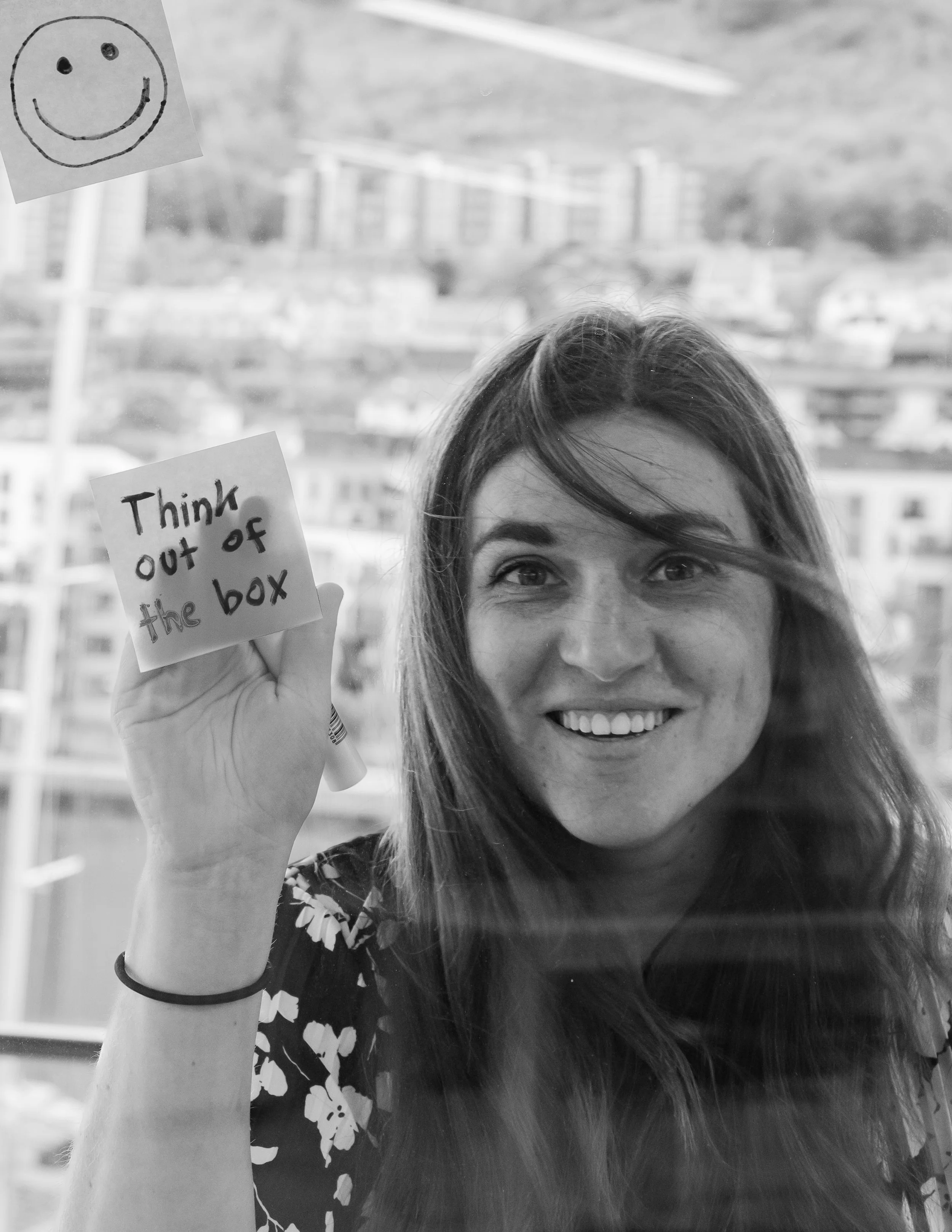 A woman holding a small note that says “think outside of the box,” standing by a window where a smiley face and the same message are taped.