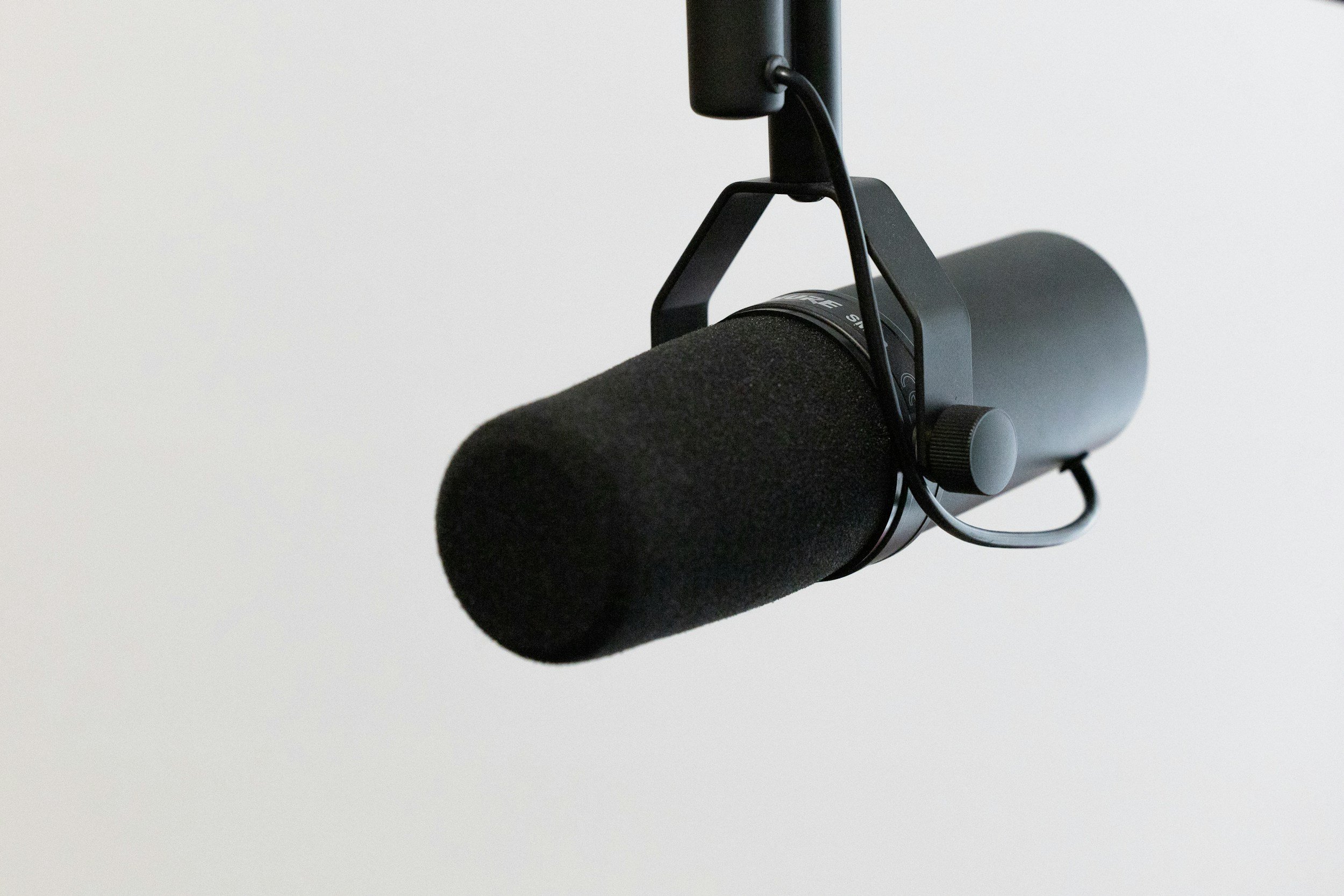 Close-up of a professional microphone with a black foam windscreen, suspended from a boom arm against a plain white background.