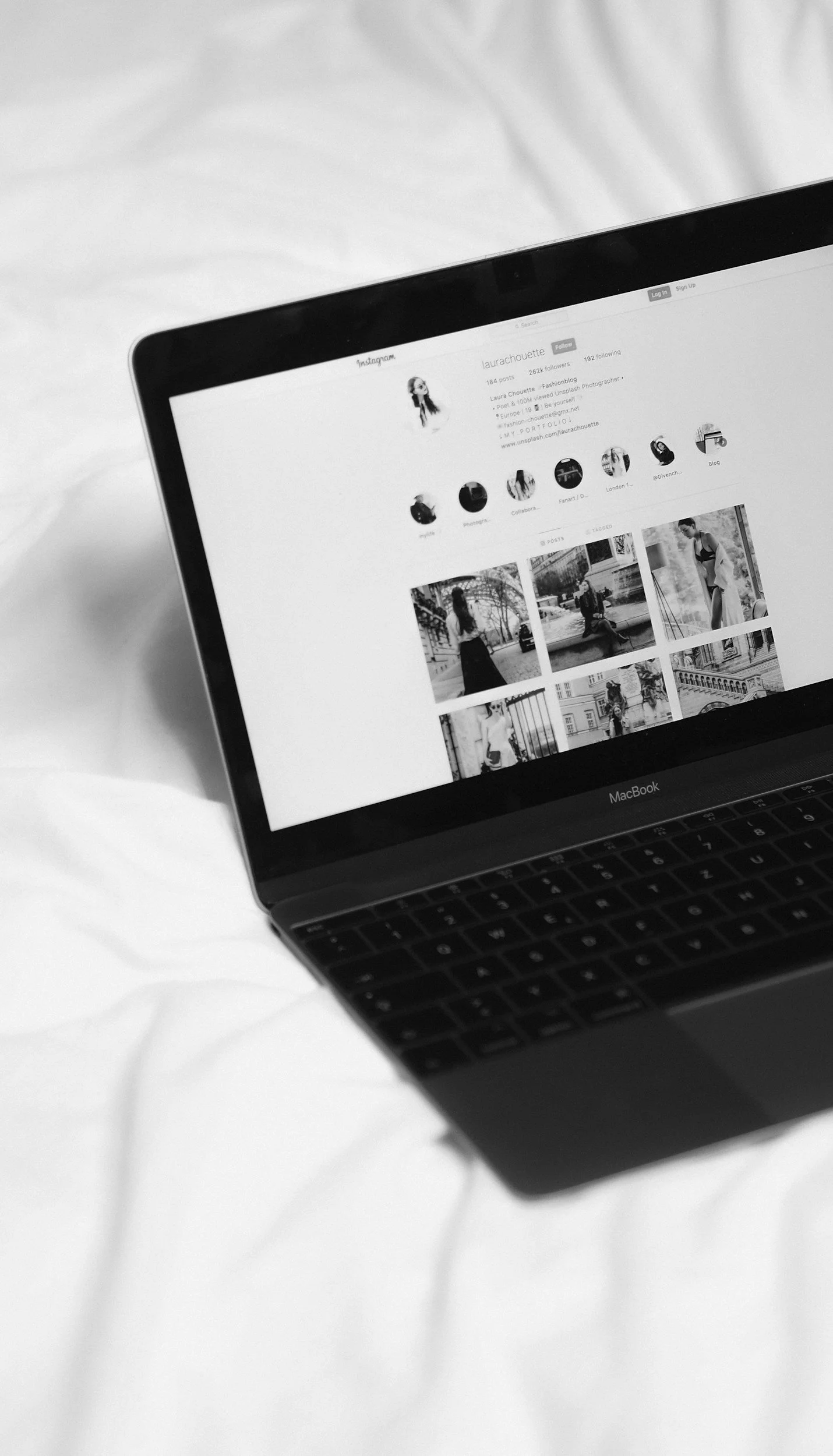A MacBook laptop displaying an Instagram profile page on a white bedsheet.