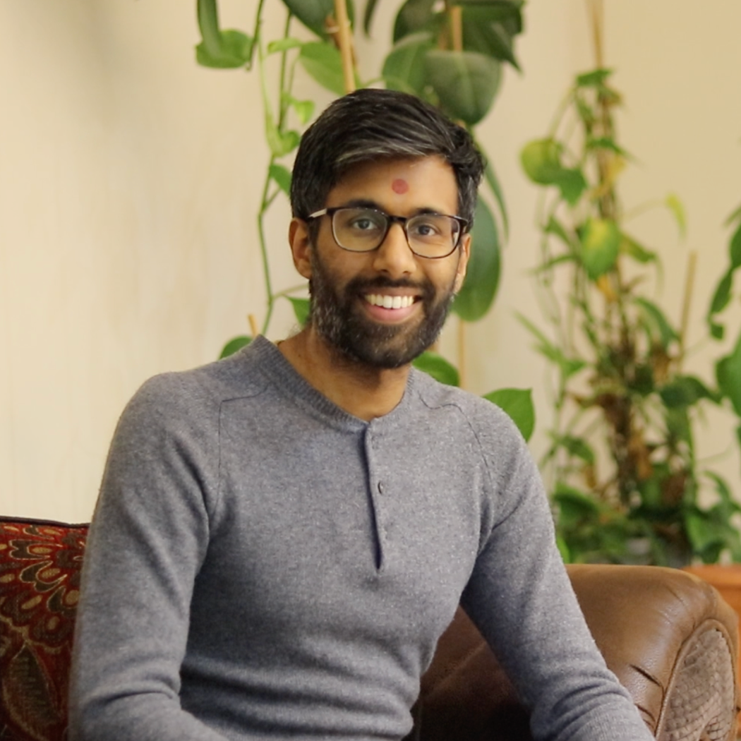A smiling man with black hair, glasses, and a beard, wearing a gray sweater, sitting on a brown leather couch, with green plants in the background.