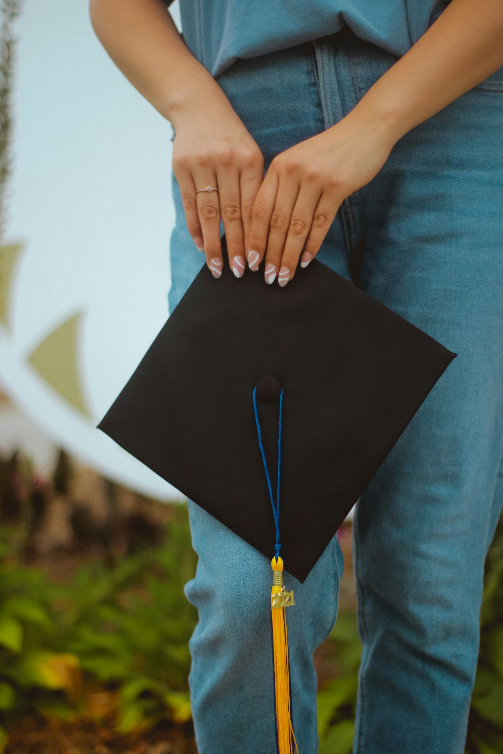 Person wearing blue jeans and a gray T-shirt, holding a black graduation cap with a yellow tassel and a blue string in an outdoor setting.