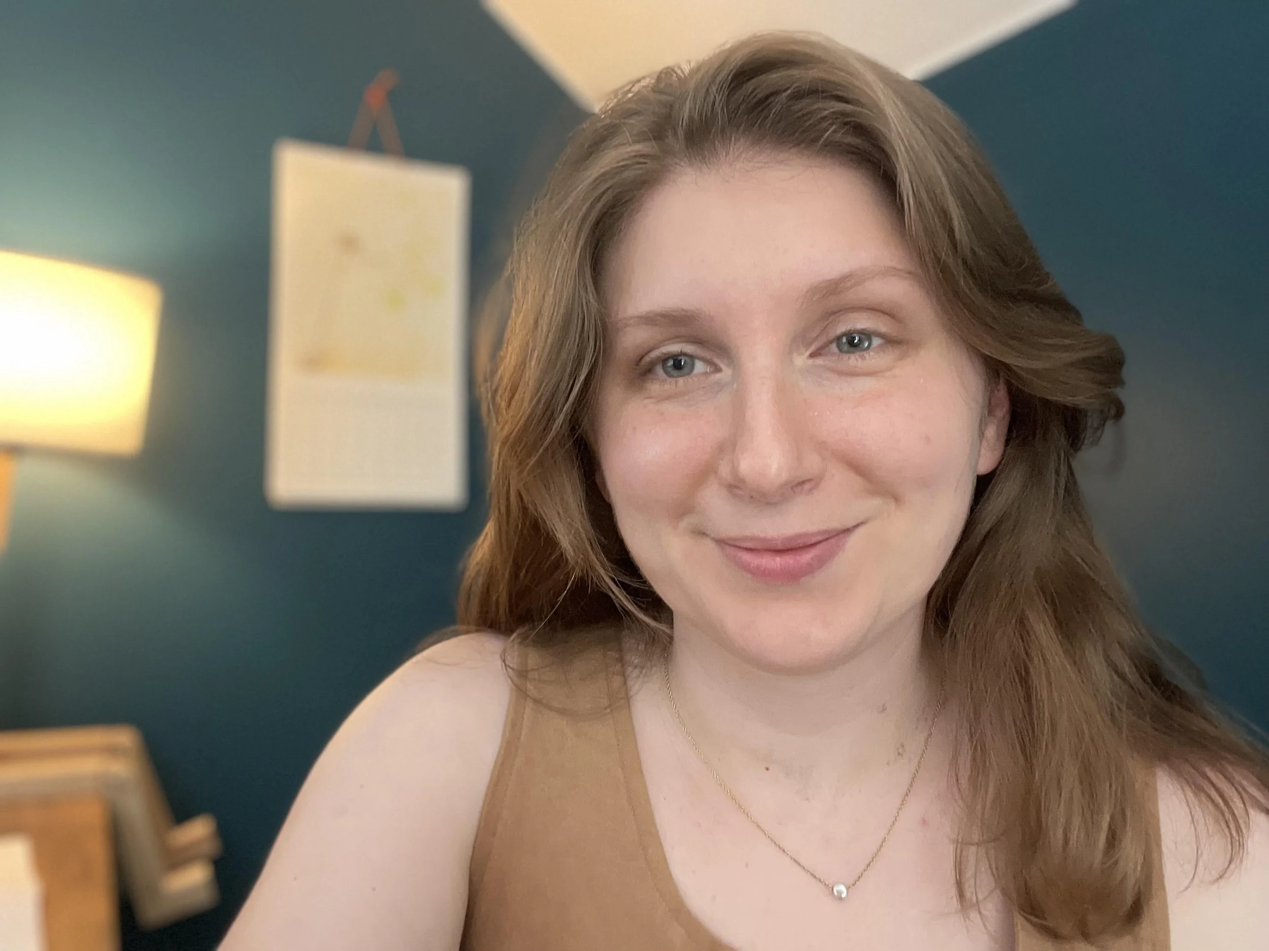 Close-up of a smiling woman with long red hair wearing a tan tank top and a delicate necklace, standing in a room with blue walls and a lit lamp.