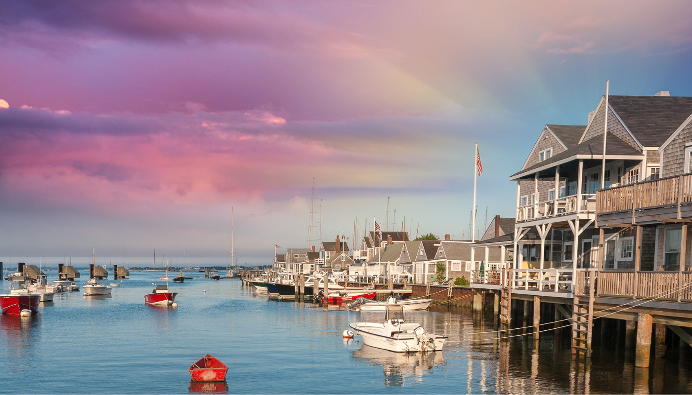 A marina with small boats docked along houses with wooden decks, under a colorful sunset sky with pink, purple, and blue clouds.