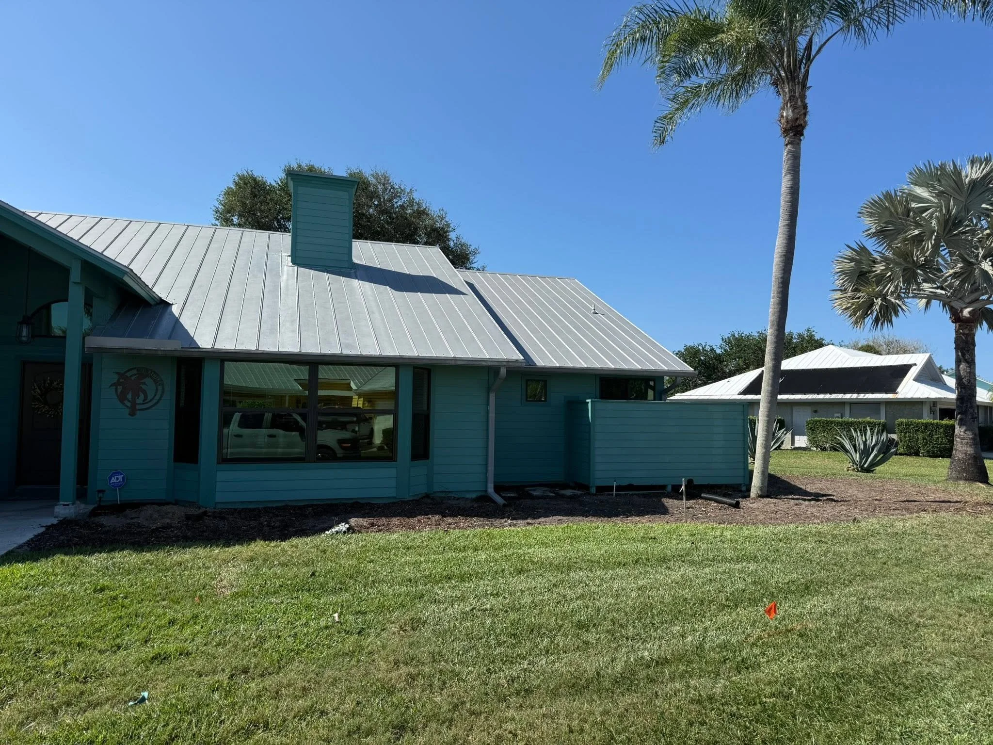 A house with a silver metal roof, a dry landscape, and palm trees under a clear blue sky.