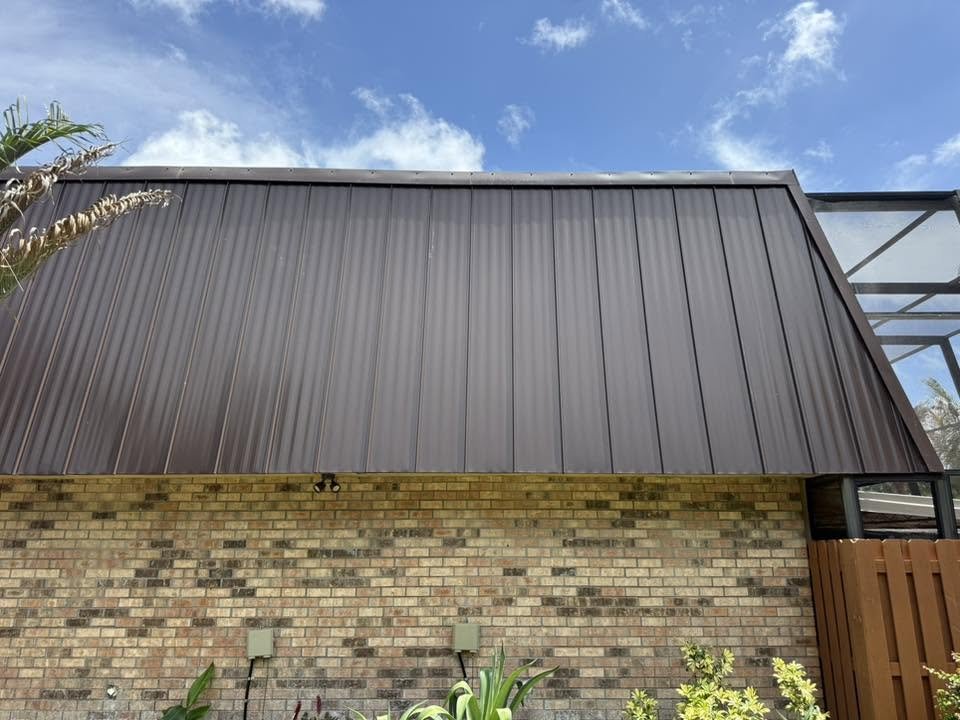 Part of a brick house with a metal roof extension, some plants, and a partly cloudy sky in the background.