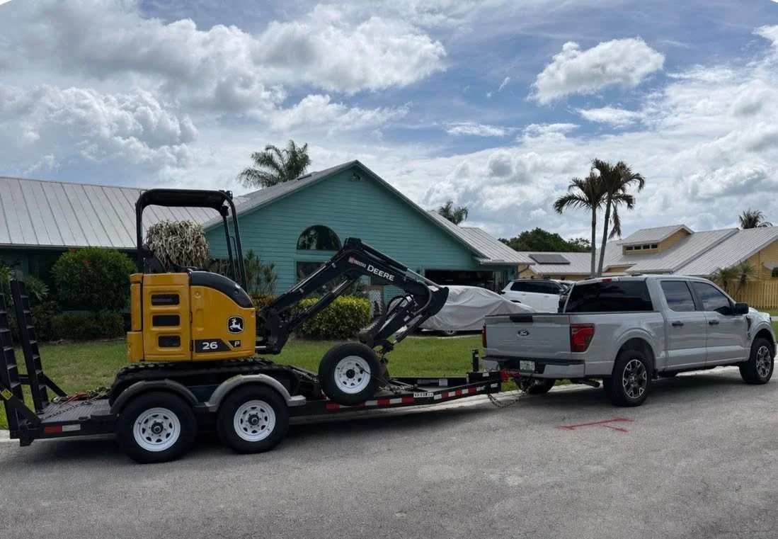 A white pickup truck parked on a residential street with a trailer carrying a small John Deere excavator. The background features a blue house, palm trees, and a partly cloudy sky.