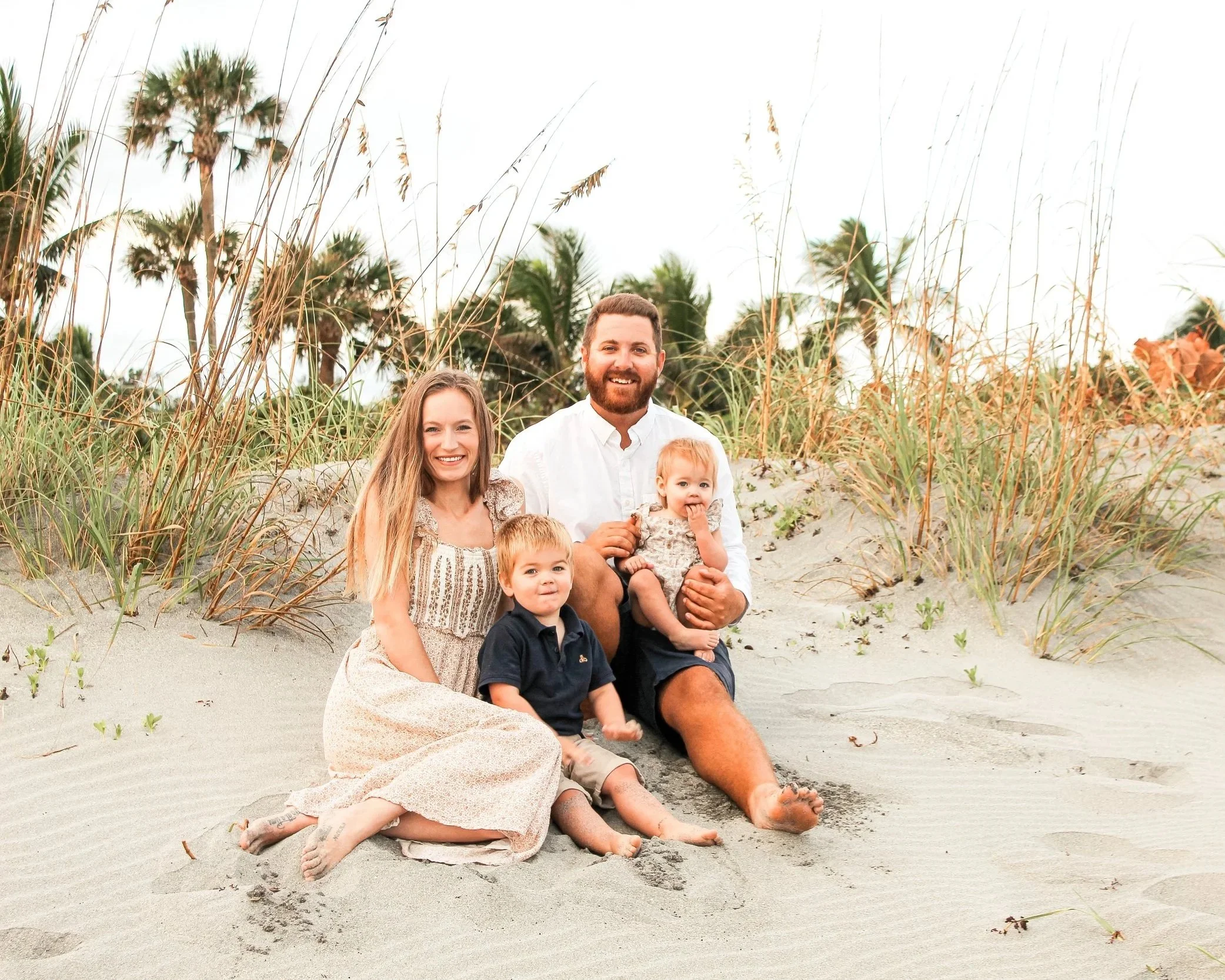 Family of six sitting on sand dunes at the beach, surrounded by beach grass and palm trees, with an overcast sky.