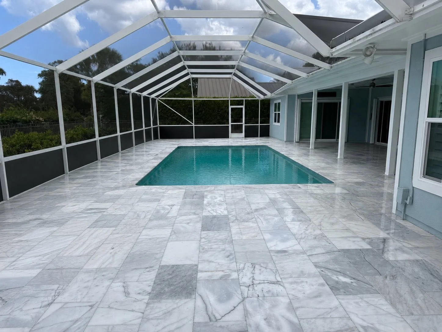 An enclosed backyard swimming pool area with marble-tiled flooring, a rectangular pool with clear blue water, and a screened structure with a sloped roof. There is a house wall with windows and a door on the right side.