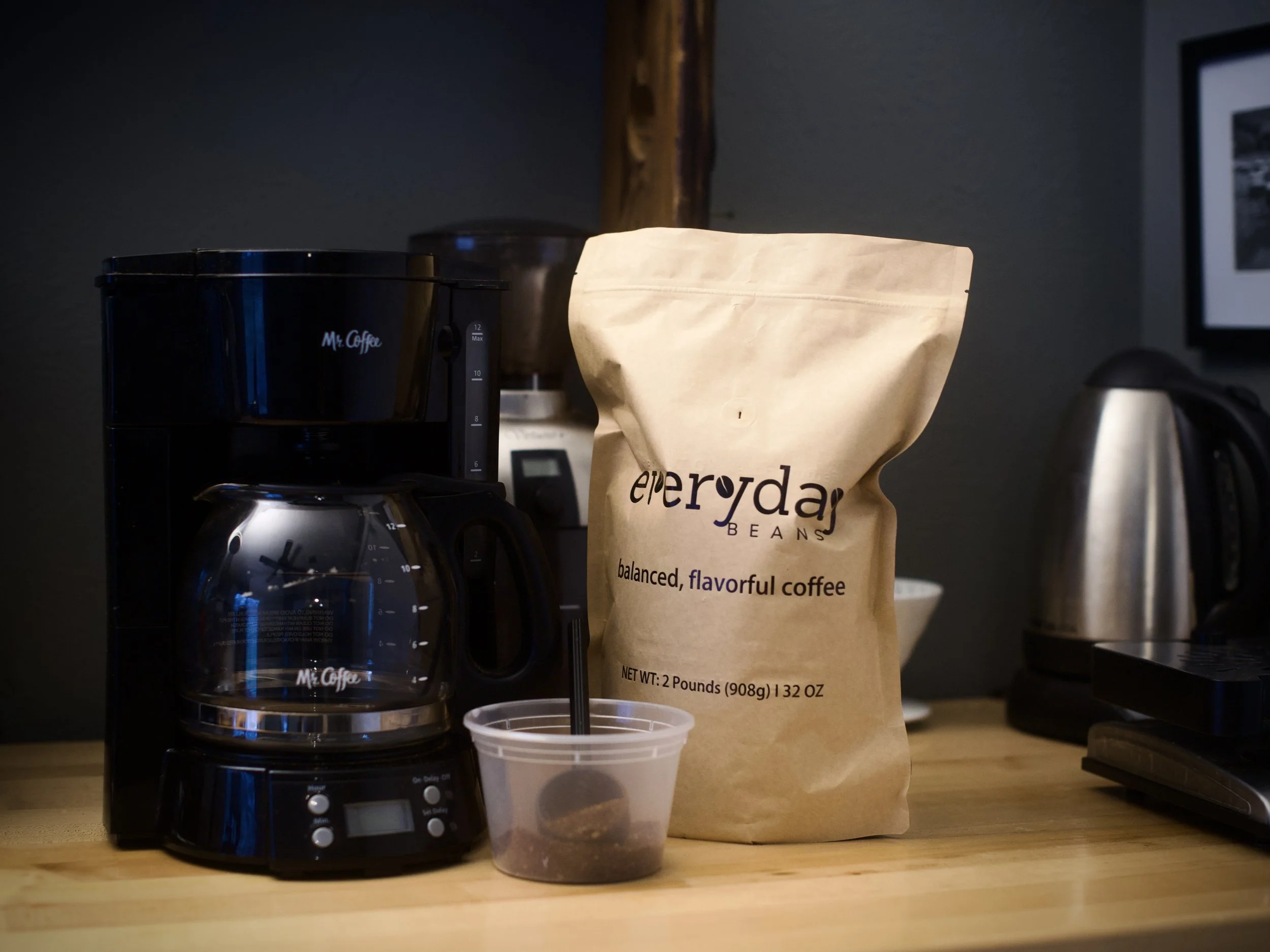 A coffee maker, a paper bag of coffee beans labeled "everyday beans," and a small plastic container with ground coffee sit on a wooden countertop in a kitchen.