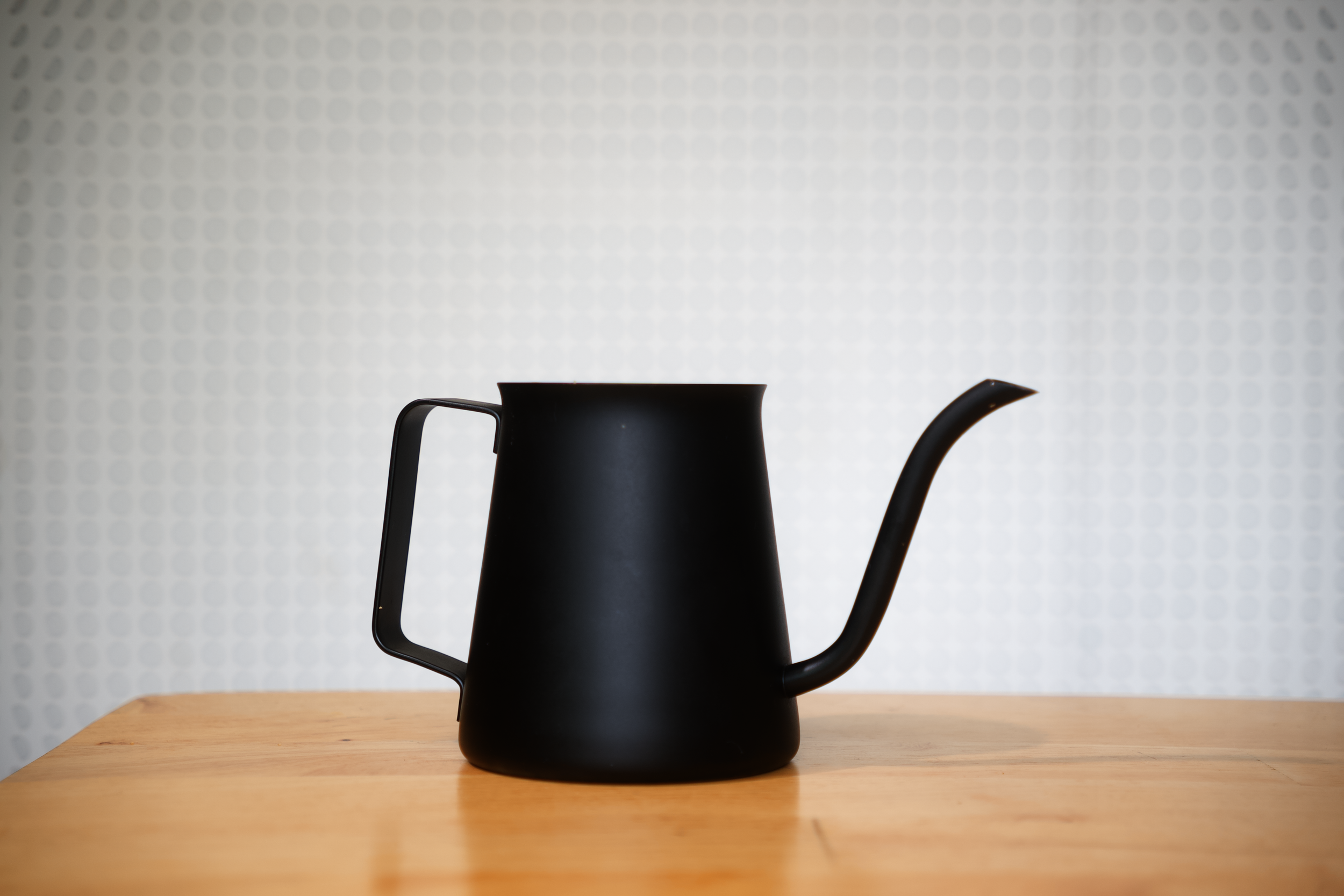 Black metal kettle with a curved spout and a rectangular handle on a wooden table, against a light dotted background.
