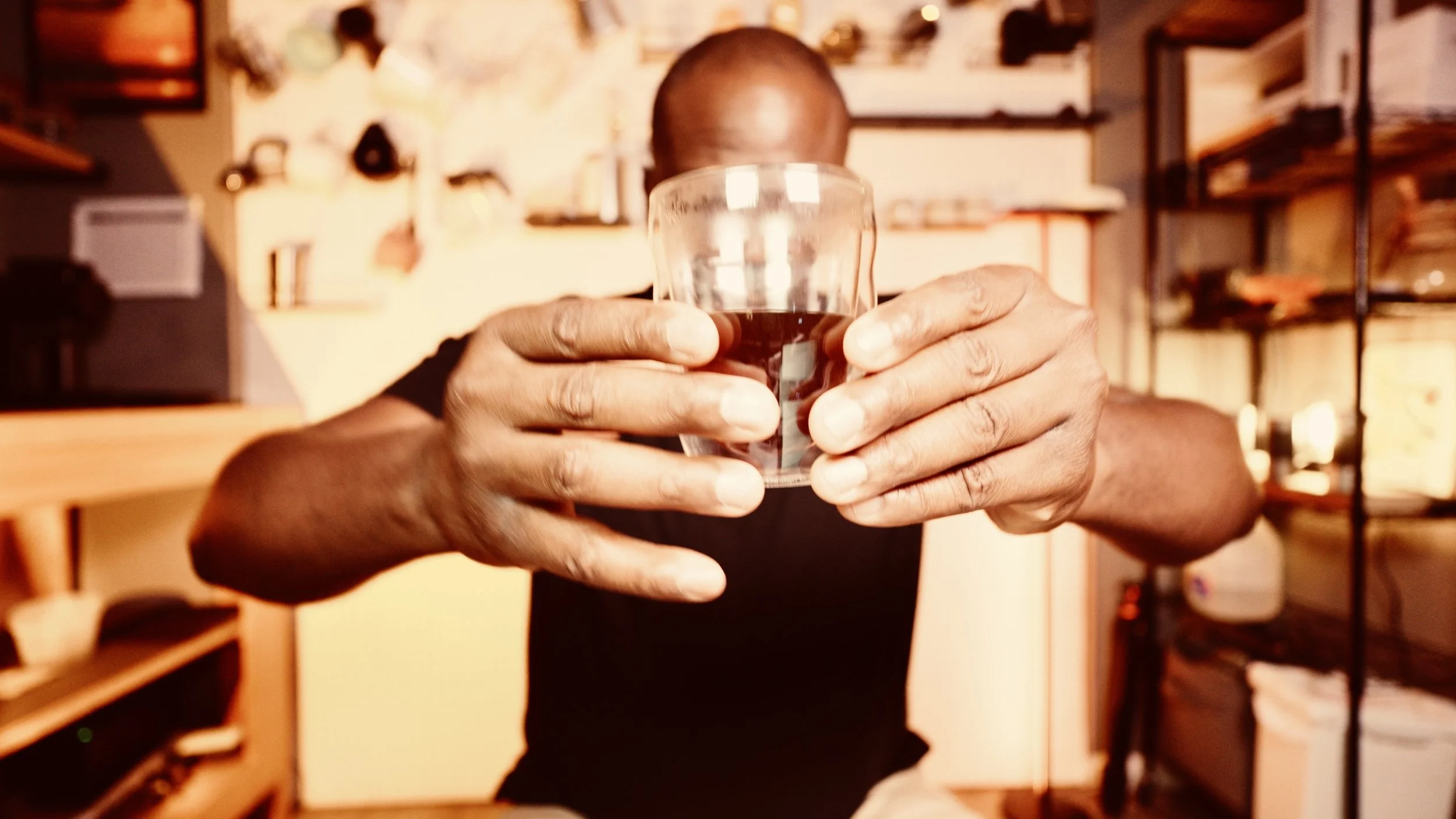 Person holding a glass of dark beverage, facing the camera in a cozy indoor setting with warm lighting and bookshelves in the background.