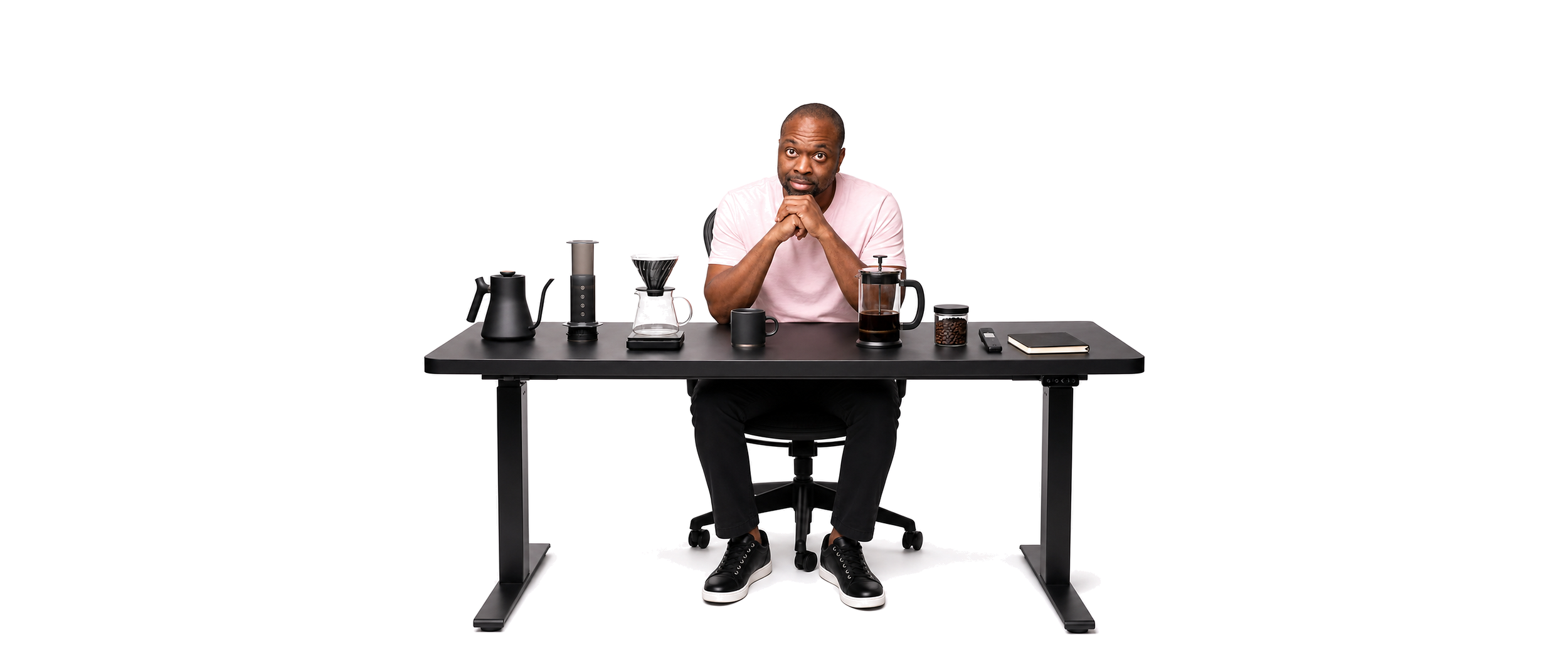 A man sitting at a black desk with various coffee-making equipment, a cup, a notebook, and a remote, against a plain white background.