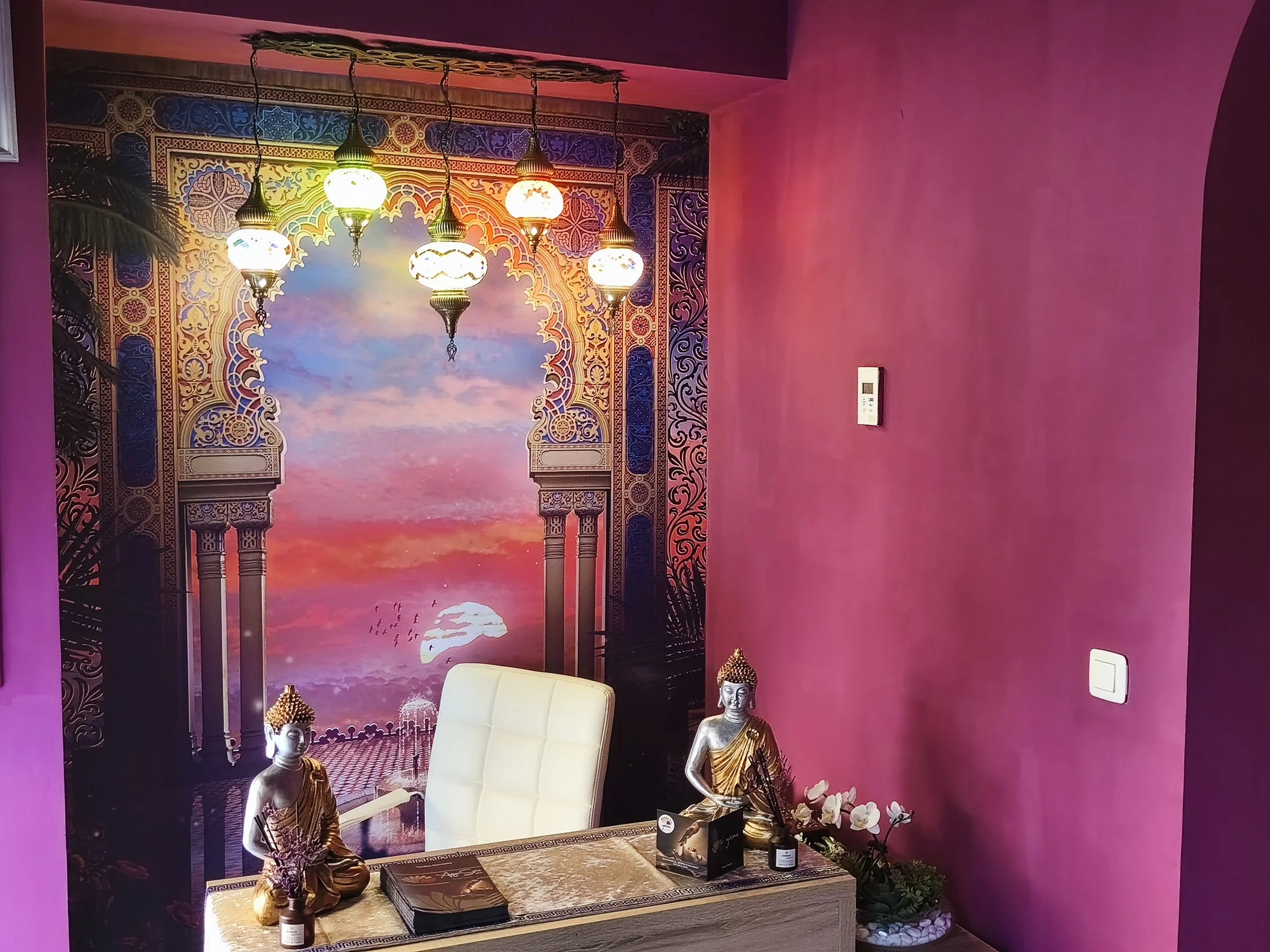 Interior de un espacio decorado con un mural que muestra un atardecer, columnas y patrones orientales. Hay una silla blanca, estatuas de Buda en color dorado, y plantas, en un ambiente con paredes rosas.