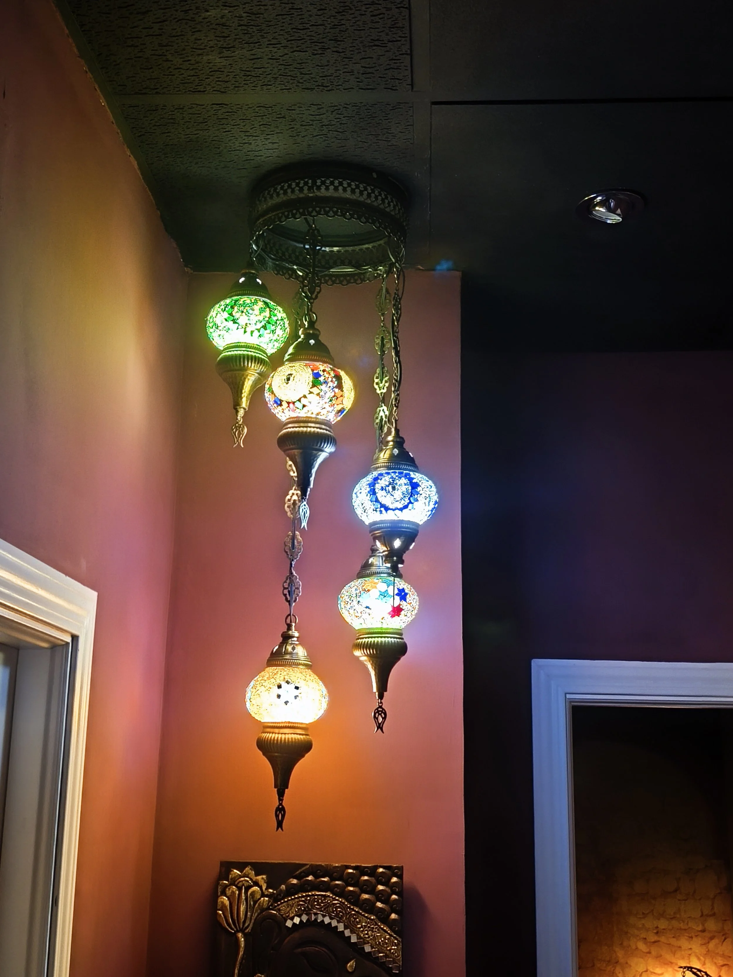 Lámparas turcas de colores brillantes cuelgan del techo en un ambiente interior con paredes en tonos cálidos y una puerta blanca con moldura decorativa.