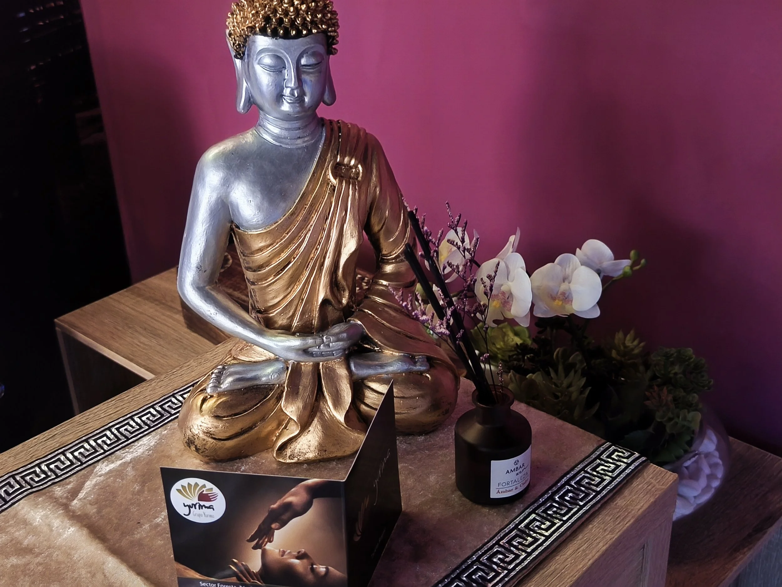 Una escultura de Buda con dorado y plateado, ubicada sobre una mesa de madera decorada con patrones griegos. Hay flores a su lado, incluyendo orquídeas blancas y otras plantas. Fondo rosa
