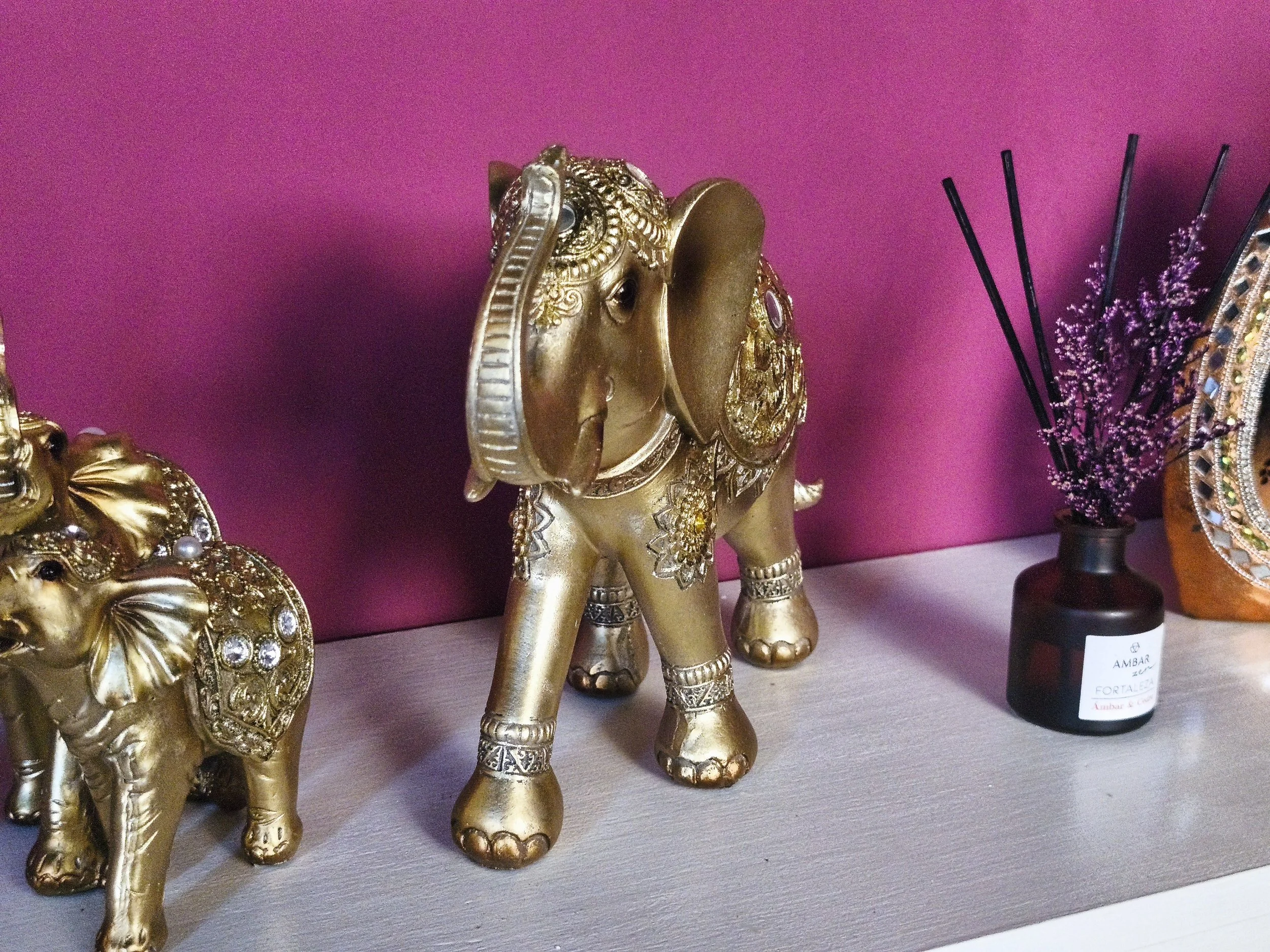 Escultura dorada de un elefante decorada con joyas y detalles ornamentales en un ambiente con pared rosa y otros objetos decorativos.