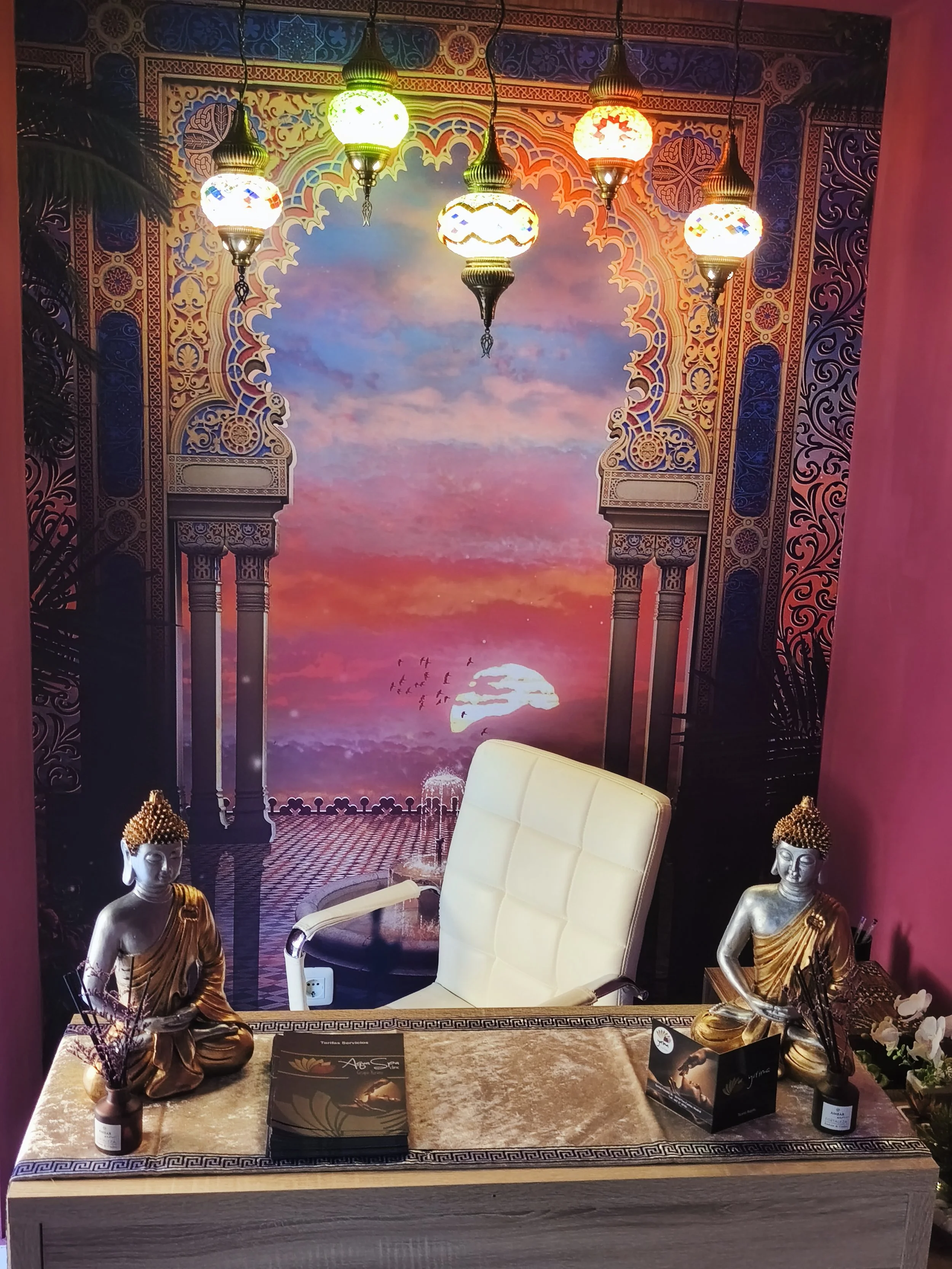 Una mesa decorada con figuras de Buda en posición de meditación, arreglos florales, y folletos. Detrás hay una pared con un mural que muestra un cielo al atardecer con un sol y aves en vuelo, rodeado de un marco arquitectónico de estilo islámico. Sob