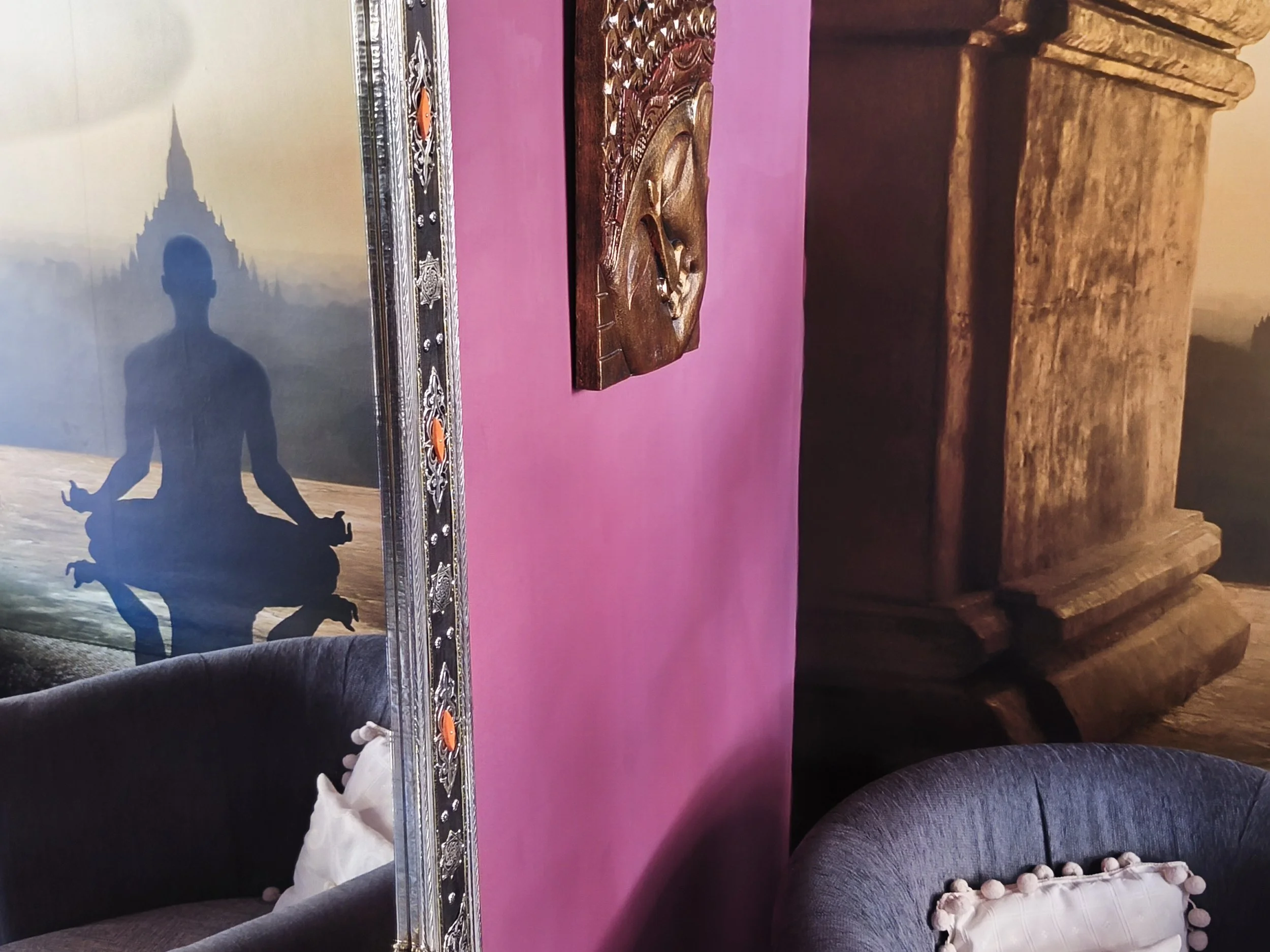 Espejo grande con marco decorativo en una pared rosa, reflejando una figura en posición de meditación y una sombra en el fondo, con muebles de madera y un sillón gris oscuro con cojines decorativos blancos y rosados.