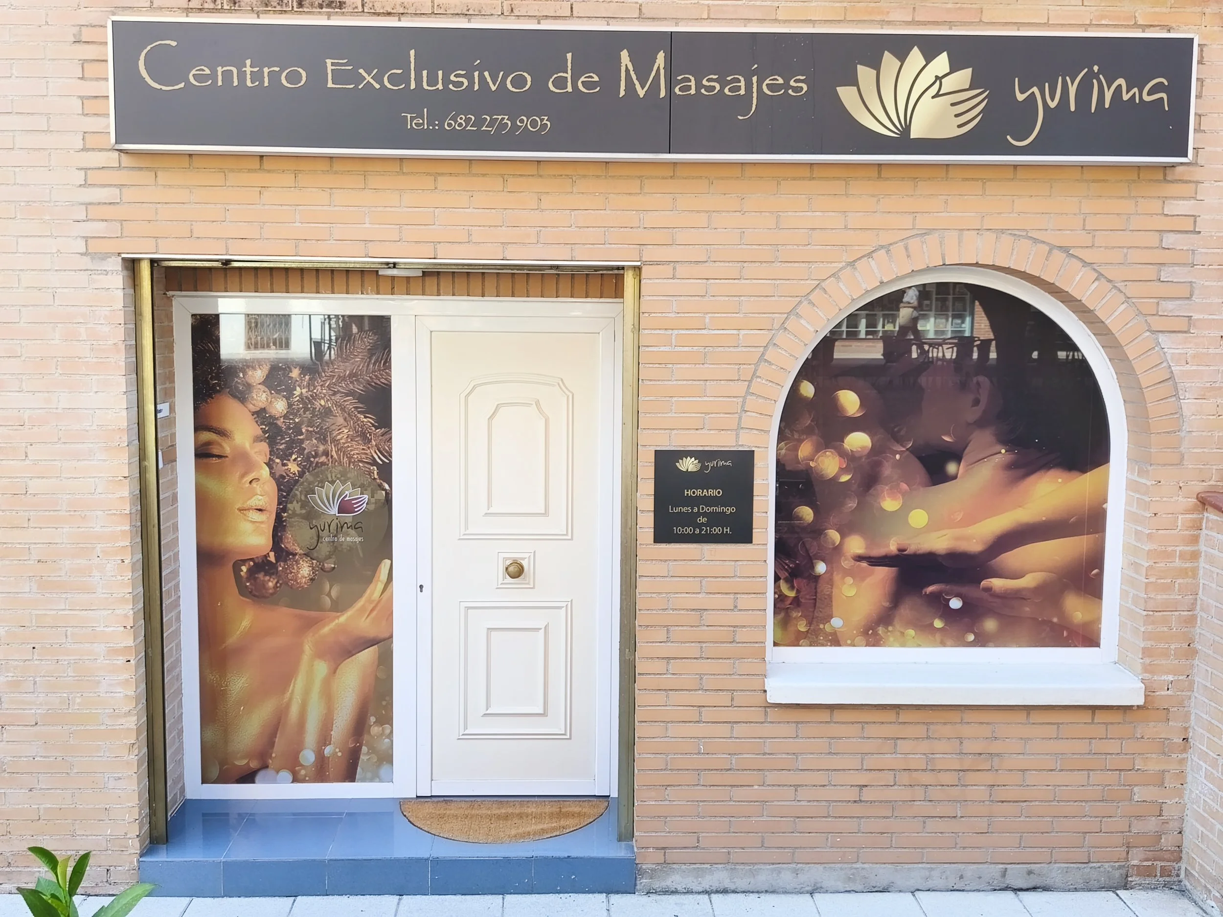 Entrada de un centro de masajes llamado 'Yurima' con un cartel en la parte superior que indica el número de teléfono y horario de atención, decorada con ventanas que muestran imágenes de personas relajadas y elementos de spa.