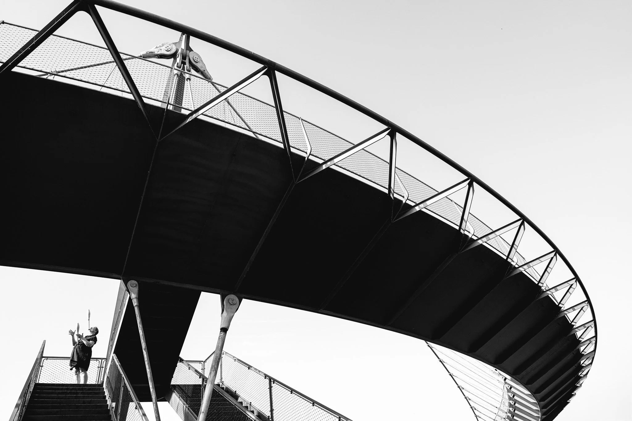 Une structure de passerelle ou escalier extérieur en métal avec une plateforme et des rampes, photographiée en noir et blanc contre un ciel clair.