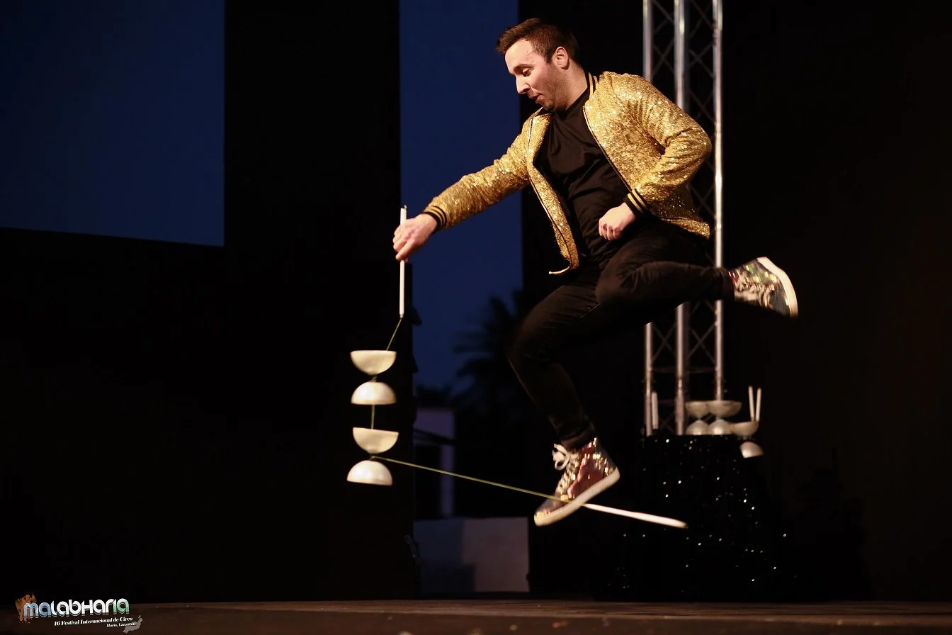 Un artiste jongle avec des assiettes en équilibre lors d'un spectacle de cirque, portant un sweat à capuche orné de paillettes dorées, sur scène sombre.