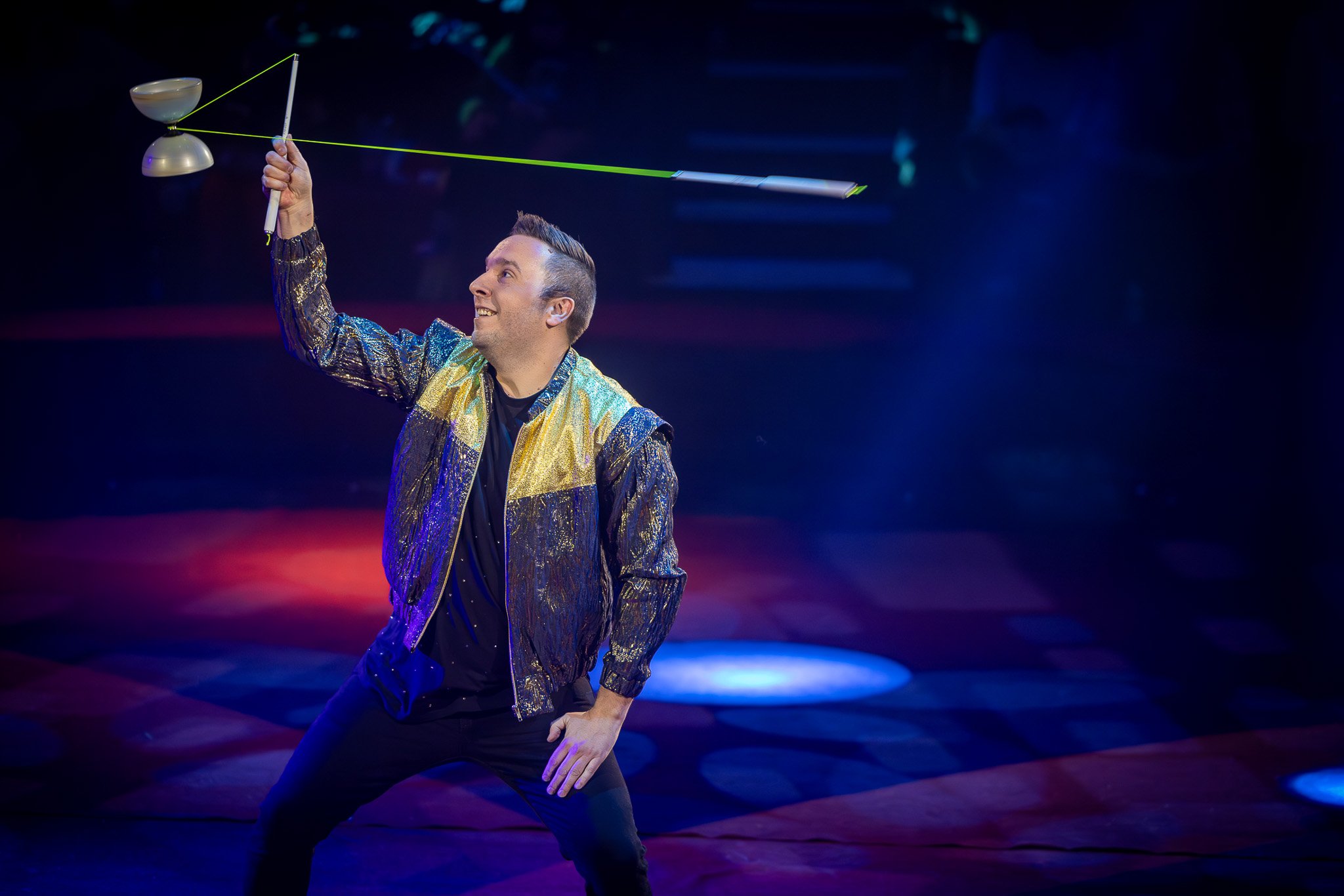 Un homme souriant en costume brillant orchestre un numéro de diabolo sur scène avec un arrière-plan sombre et éclairé par des lumières colorées.