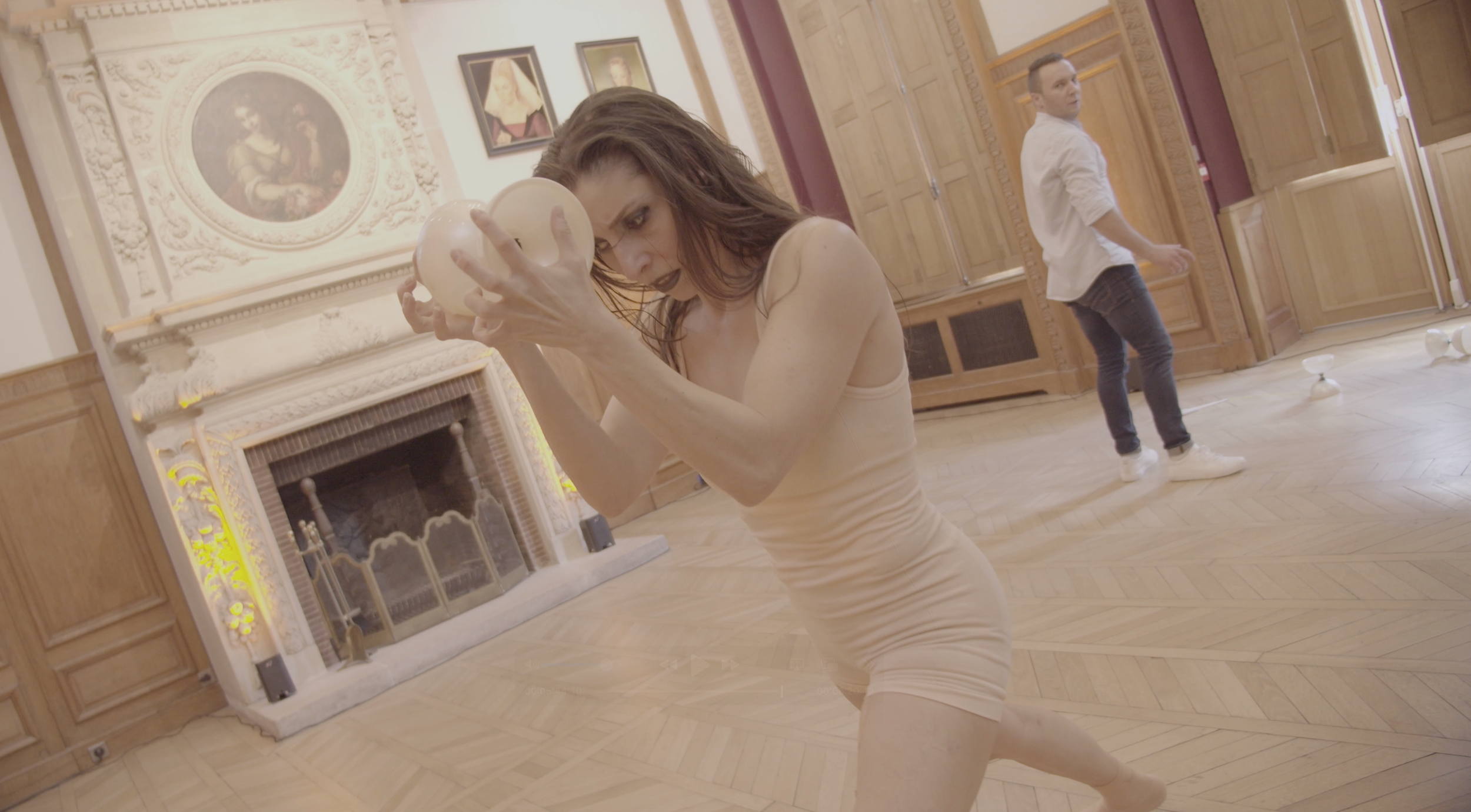 Une femme dans une salle d'art, apparemment en mouvement ou en danse, tenant une sculpture ou un objet rond, avec un homme au fond qui regarde dans une autre direction. La salle a des murs en bois et des œuvres d'art accrochées.