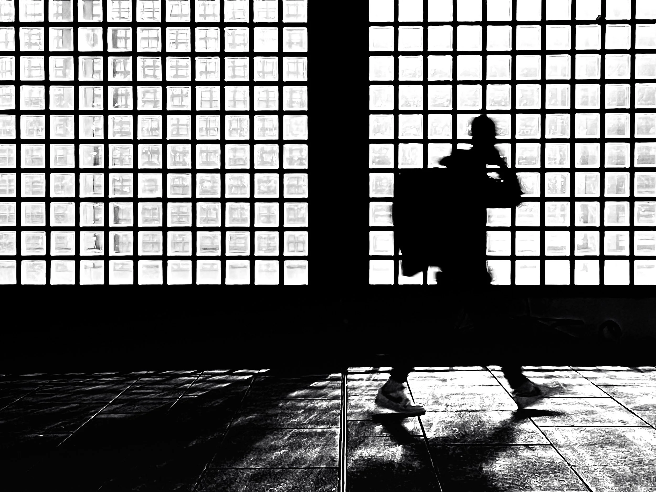 Silhouette d'une personne en train de marcher devant une grande fenêtre en verre. La lumière venant de l'extérieur crée un contraste élevé, rendant la personne presque noire dans l'image.