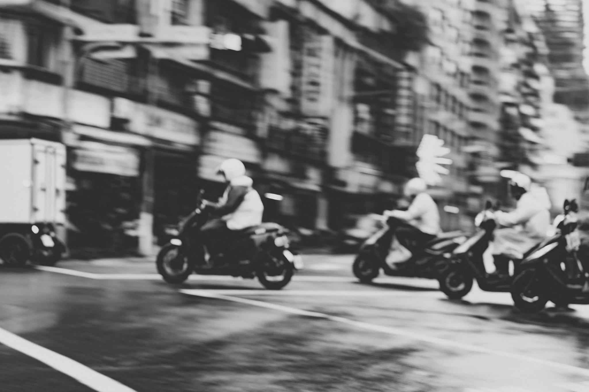 Une file de motos roulant sur une route urbaine en noir et blanc, avec des immeubles en arrière-plan.