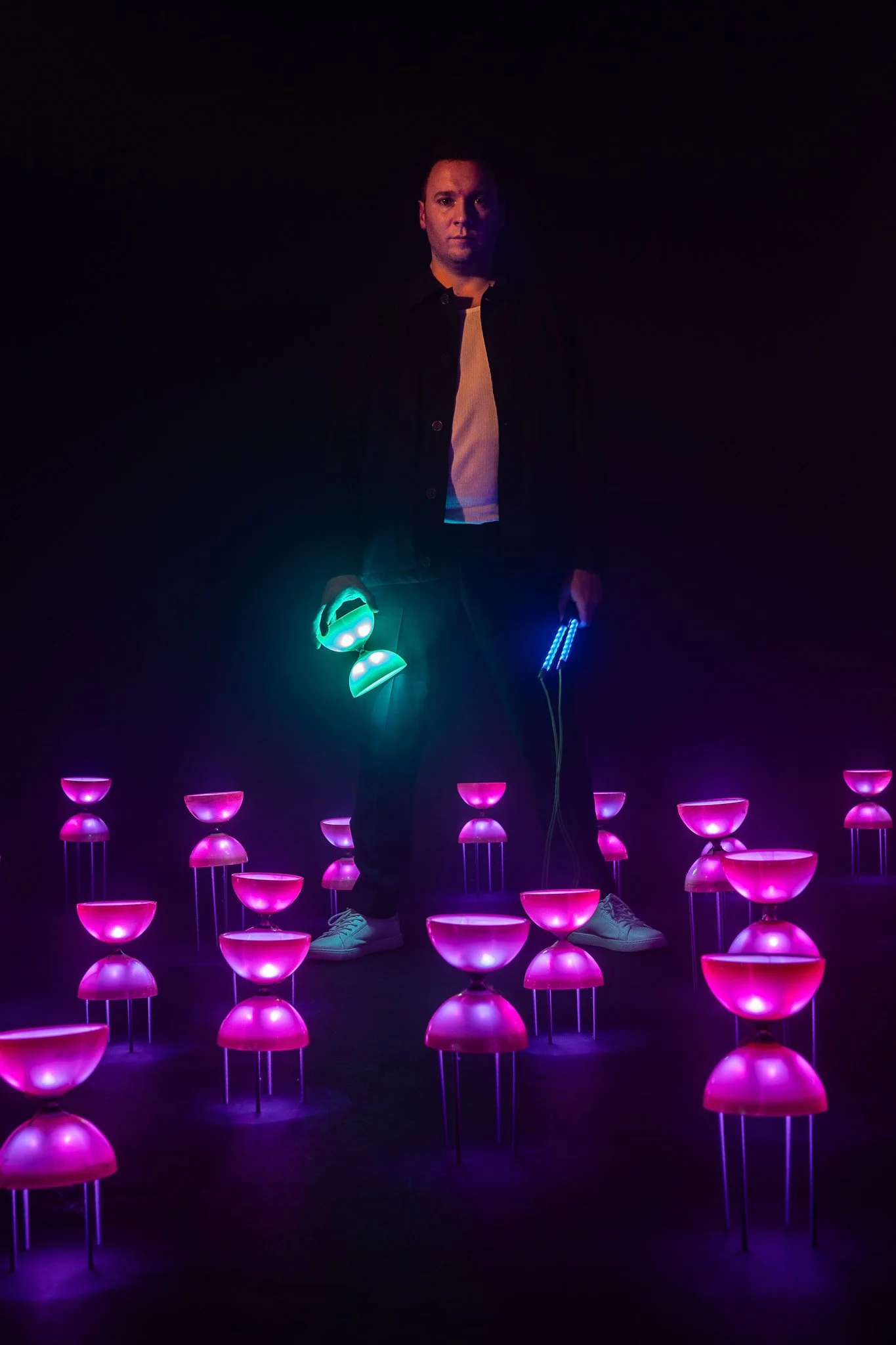 Un homme debout dans une pièce sombre, entouré de nombreuses lampes colorées en forme de bol, émettant des lumières roses, vertes et bleues, créant une ambiance futuriste.