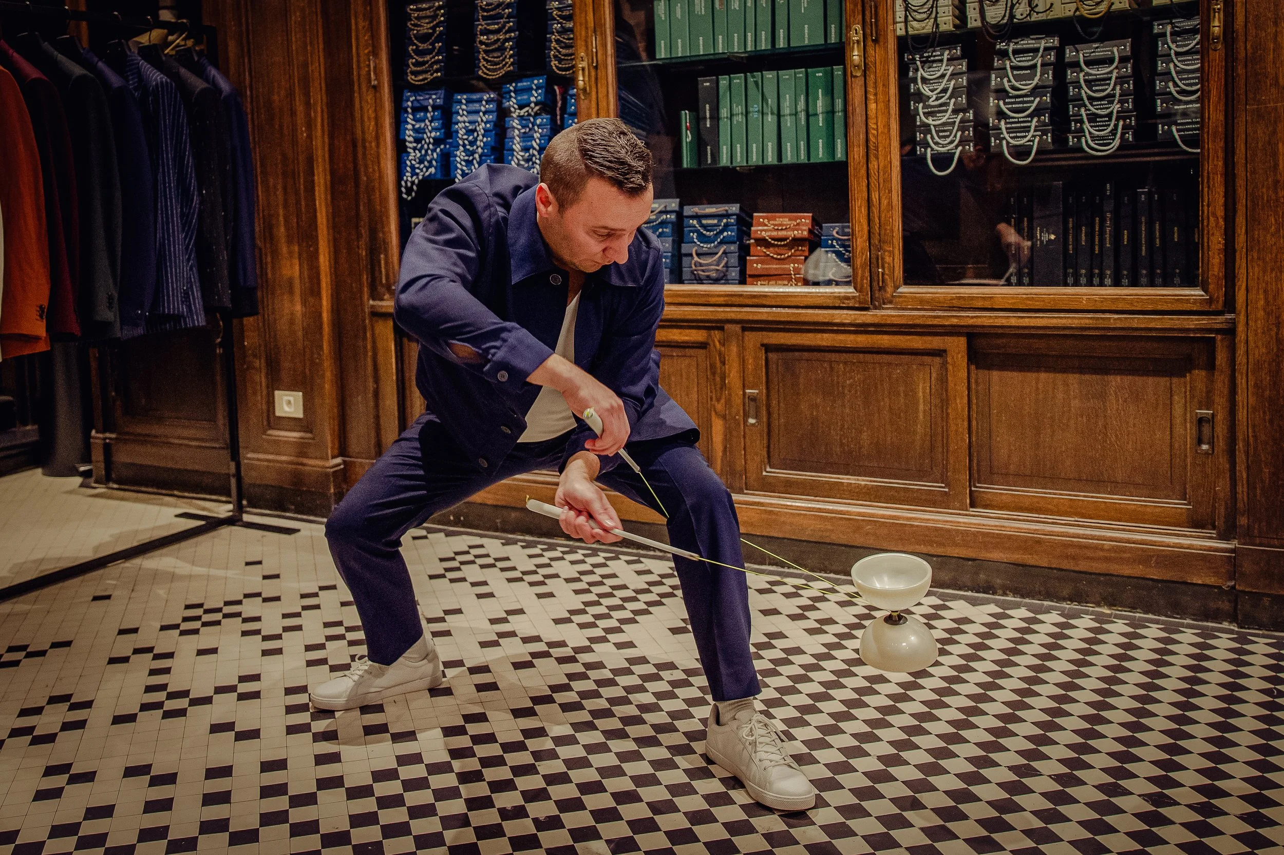 Un homme joue au billard dans une pièce avec des murs en bois et des étagères remplies de livres et de bijoux, portant une veste bleue, une chemise blanche et des baskets blanches, en position accroupie en train de tirer sur une queue de billard.