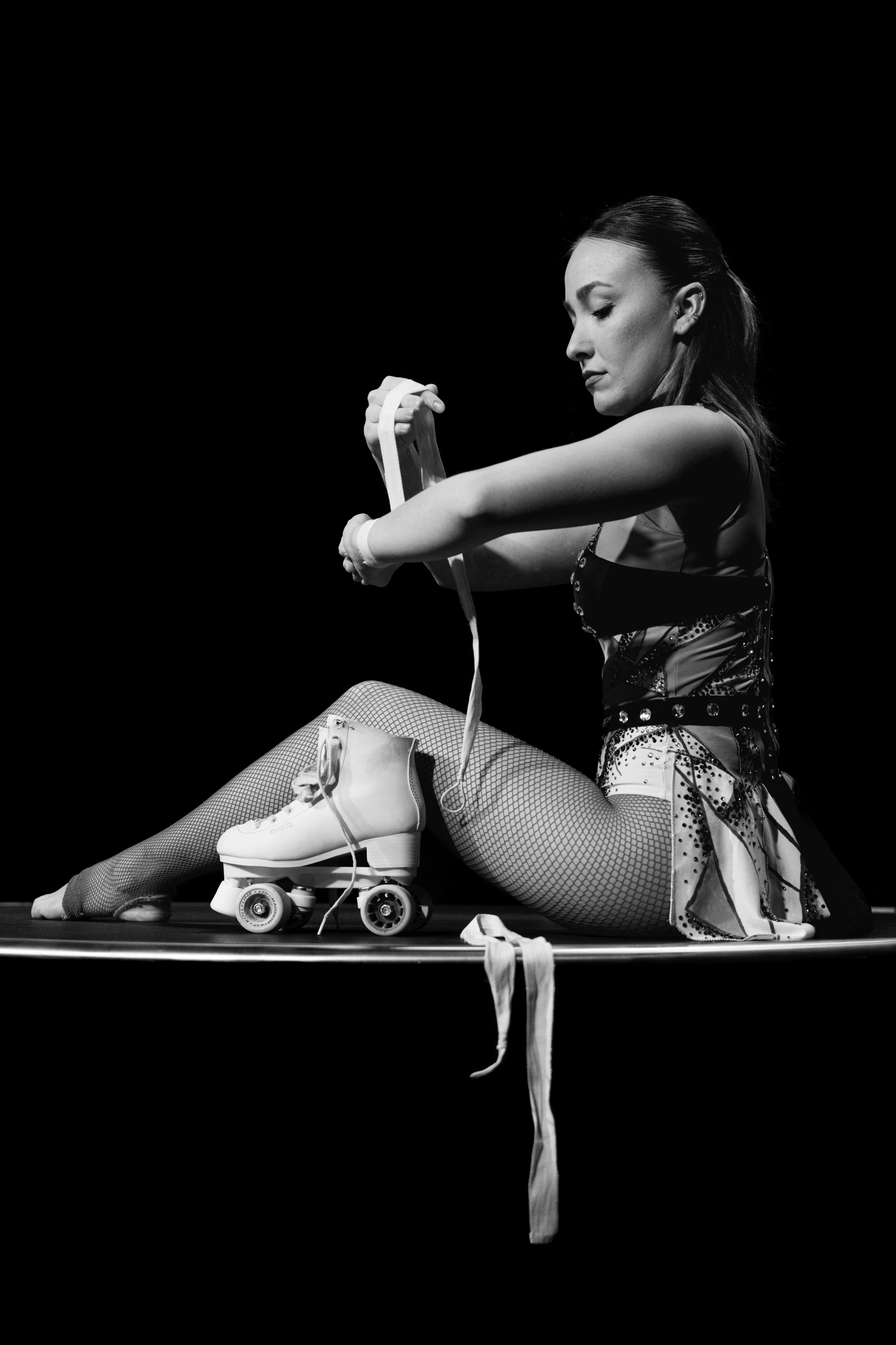 Une femme en patins à roulettes et costume de danse, assise sur un tapis noir, en train d'enrouler une bande de tissu, avec des rollers à proximité, dans un décor sombre.