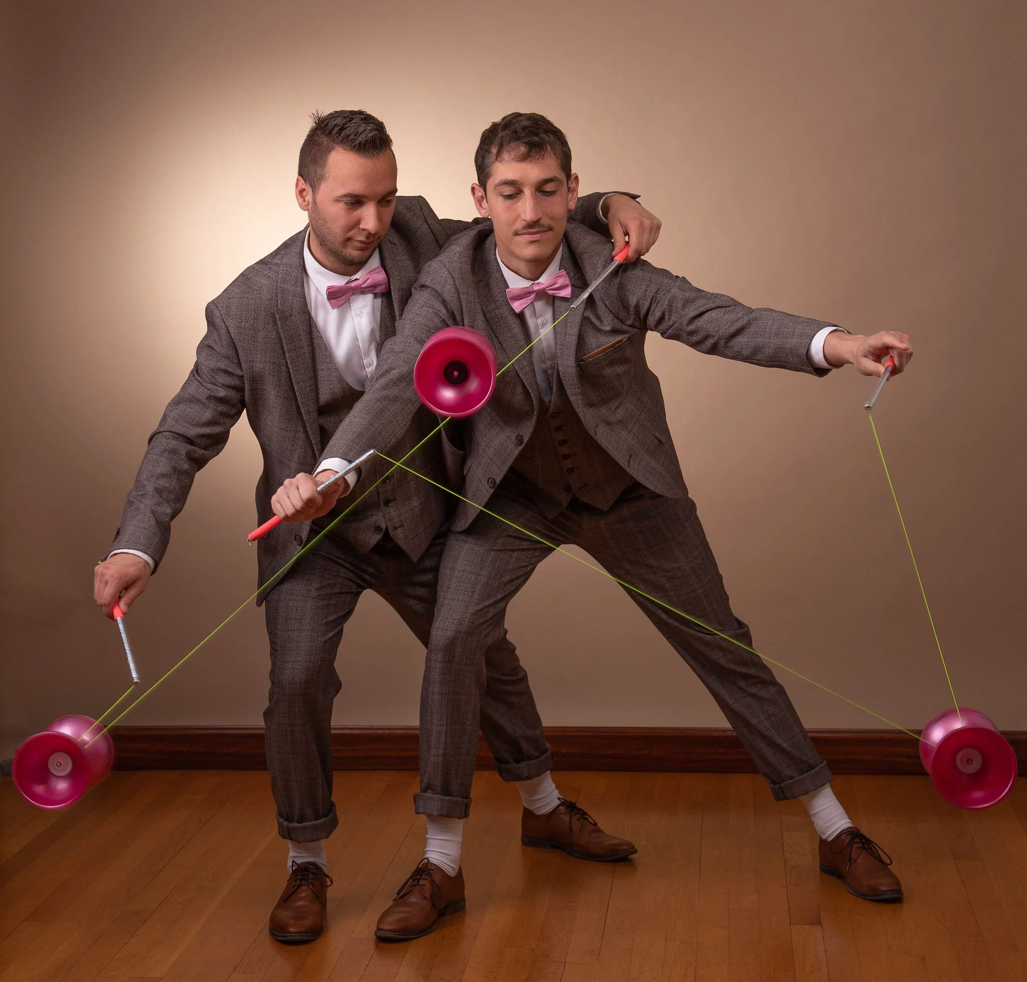 Deux hommes habillés formellement en costume avec nœuds papillons roses dans une salle avec un sol en bois clair, manipulant une ficelle jaune et entourés de ballons roses, one d'eux se doubant d'une megaphone rose.