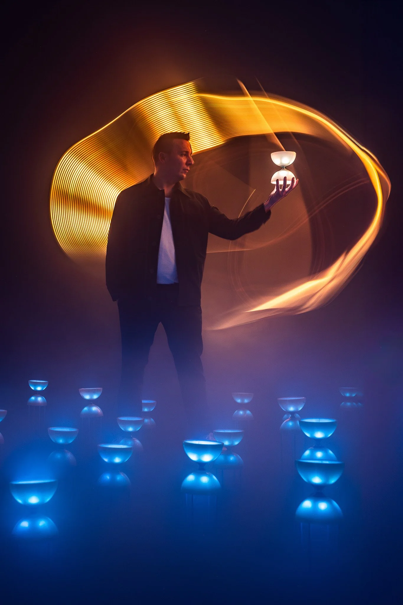 Un homme debout dans une pièce sombre, tenant une coupe à la main, entouré de lumière orange en formes de lignes fluides, avec des bols lumineux bleus au sol.